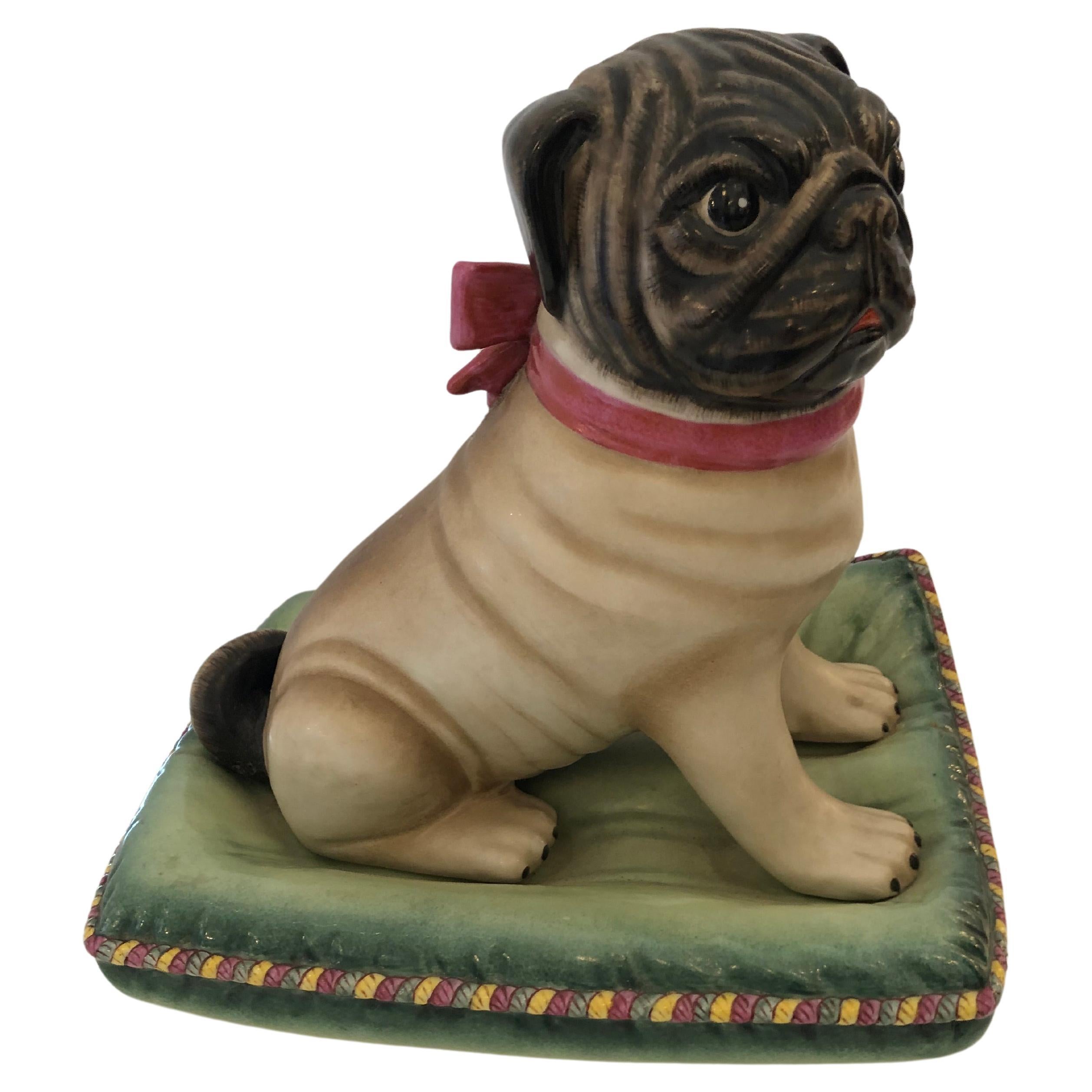 Adorable perro carlino de cerámica pintada posado sobre un suntuoso cojín.  Los colores son negro apagado, crema, verde y rosa.  El lazo alrededor del cuello del perrito es un toque perfecto.