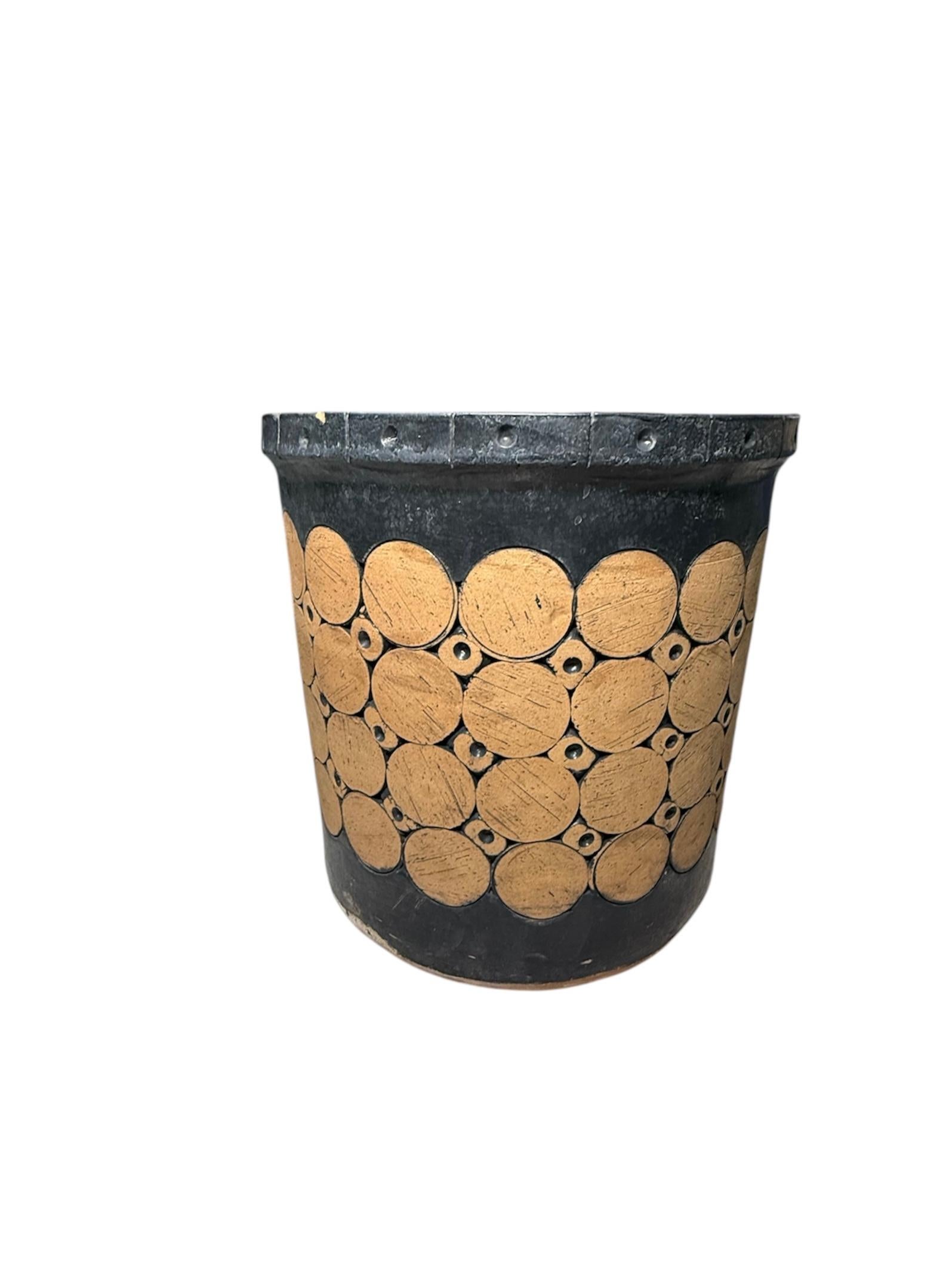 This is a Isla del Sol Puerto Rican Stoneware Round Planter. It depicts a heavy black and brick color planter. It is adorned with four rows of large circles alternated by three rows of small circles enhanced by a black color central concavity. Its