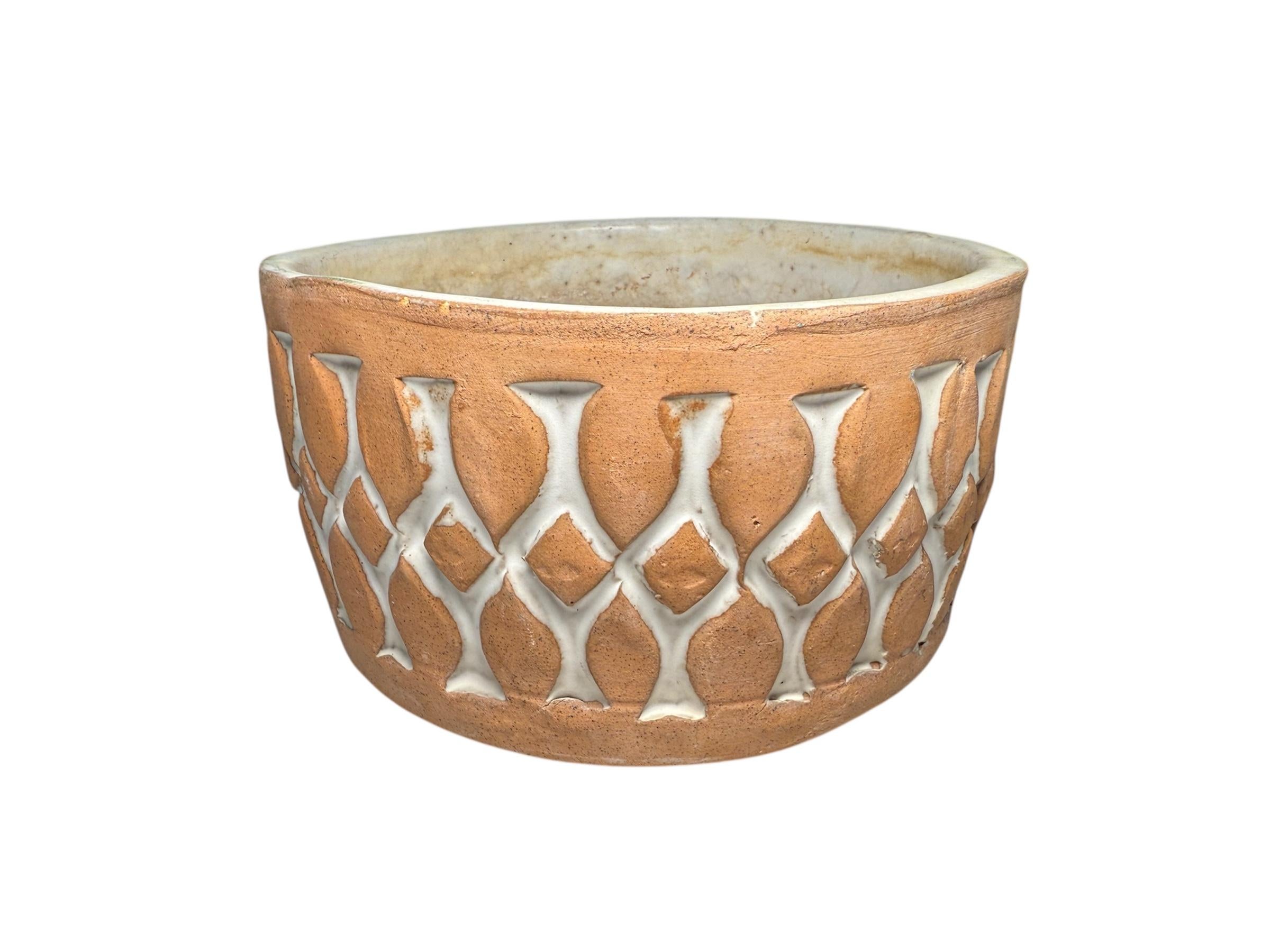 This is an Isla del Sol Puerto Rican Stoneware Planter. It depicts a round small bowl  with a pearly white background and decorated with two rows of brick color leaves and in the center of them, a row of brick color triangles. The upper and lower