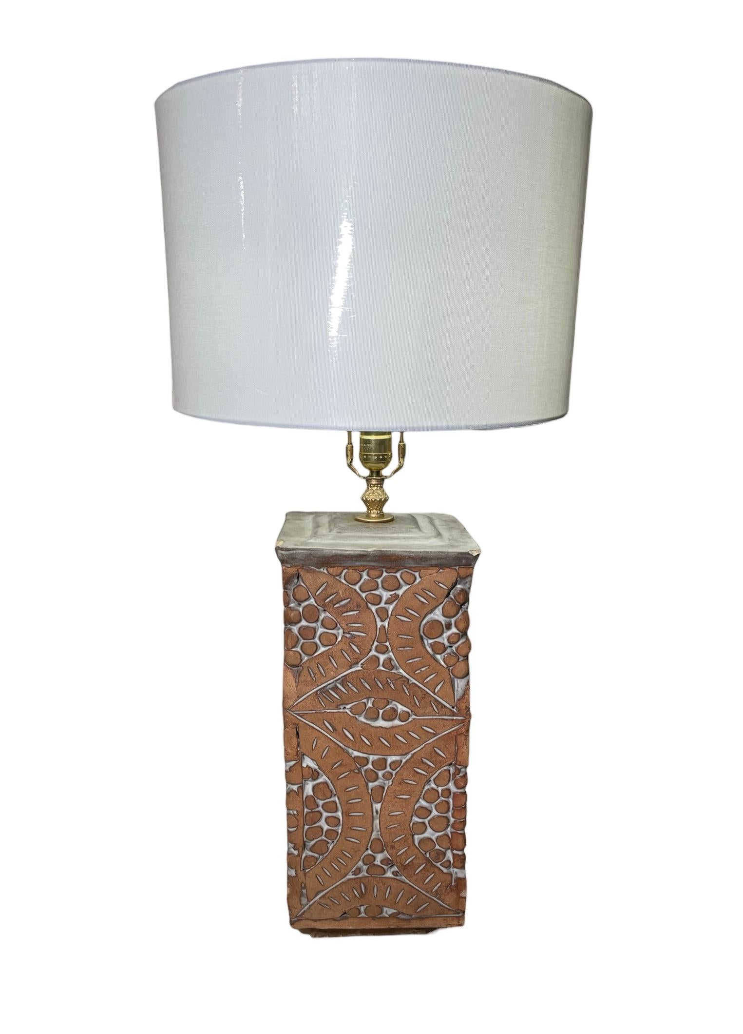 This is an Isla Del Sol Stoneware Lamp. It depicts a table lamp with a rectangular stoneware base decorated with a relief pattern of brick color leaves and folded leaves. The center of the leaves are adorned with a relief of asymmetrical brick color
