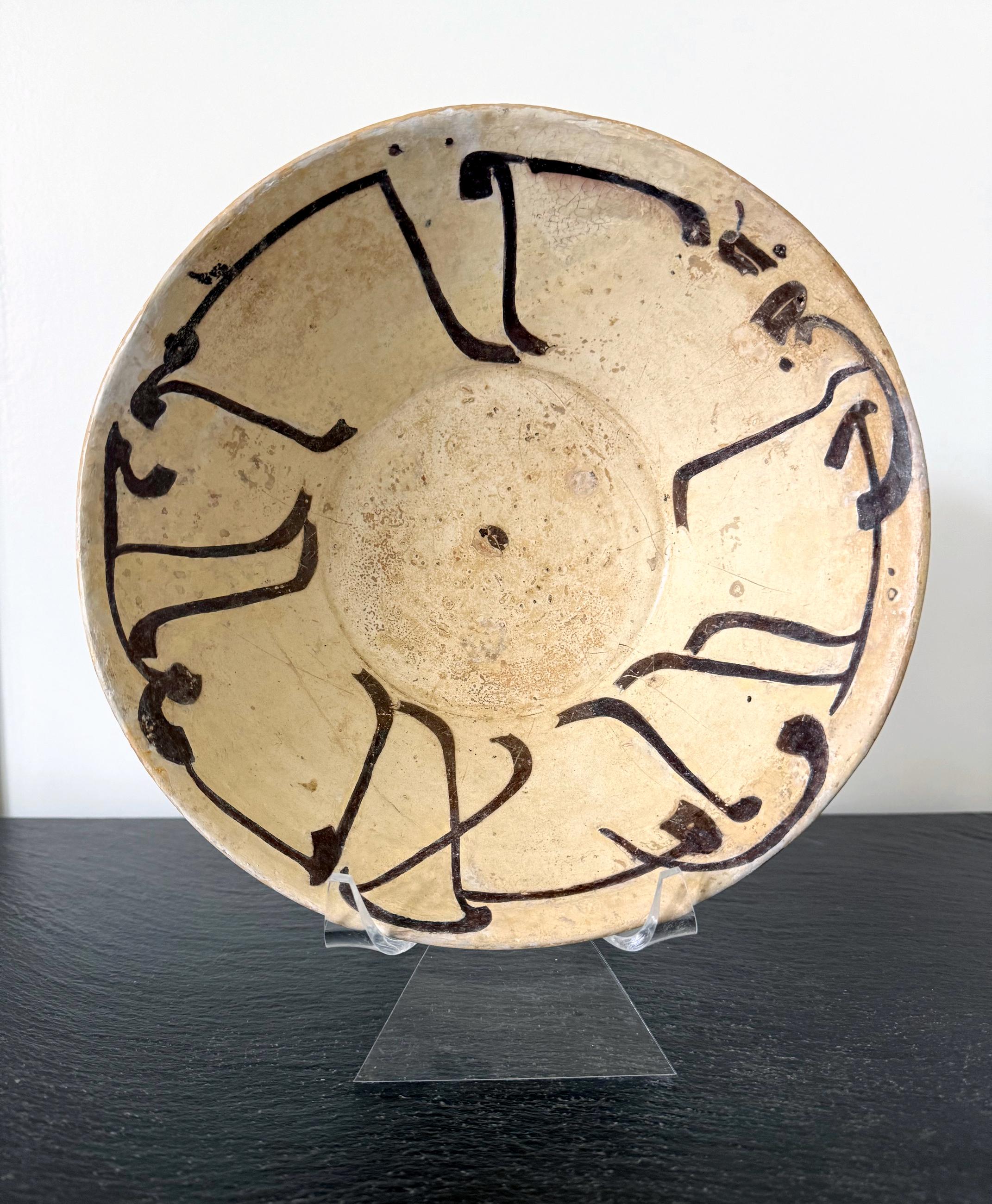 Cuenco de barro cocido de forma cónica sostenido por un anillo de pie corto cubierto de engobe blanco y decorado con pintura en engobe negra de caligrafía en escritura cúfica. Este tipo de cuenco se identifica como cerámica de Nishapur, hacia los
