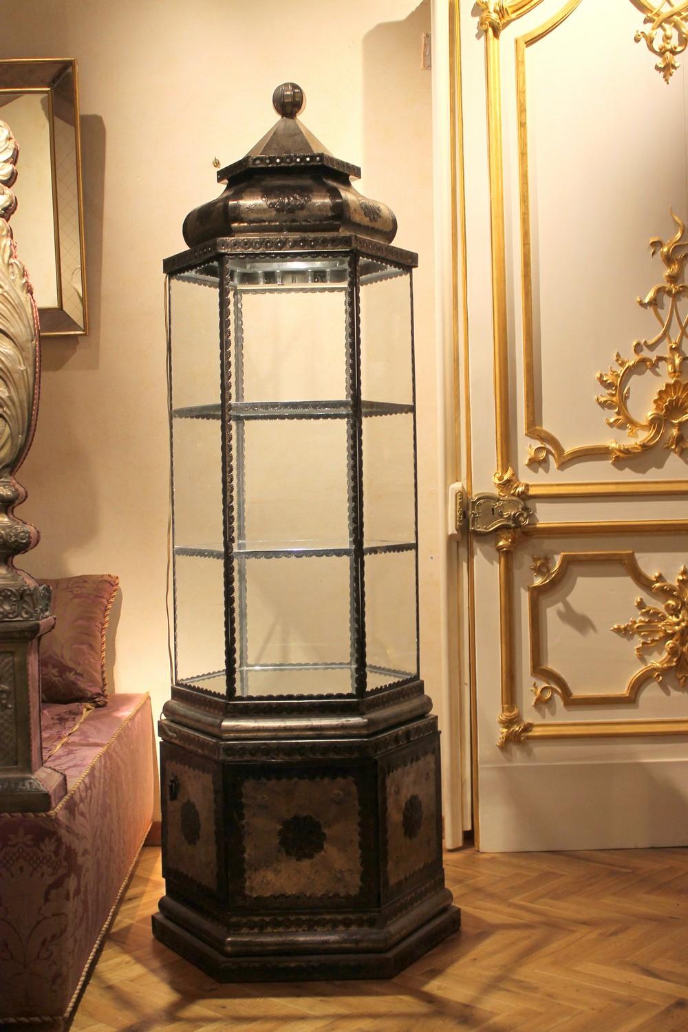 Unique of its kind, this tall hexagonal display case recalls a Middle Eastern artisan and artistic workmanship both in its Islamic style architectural shape and metal embossed decorations. This whimsical vitrine cabinet could be dated between the