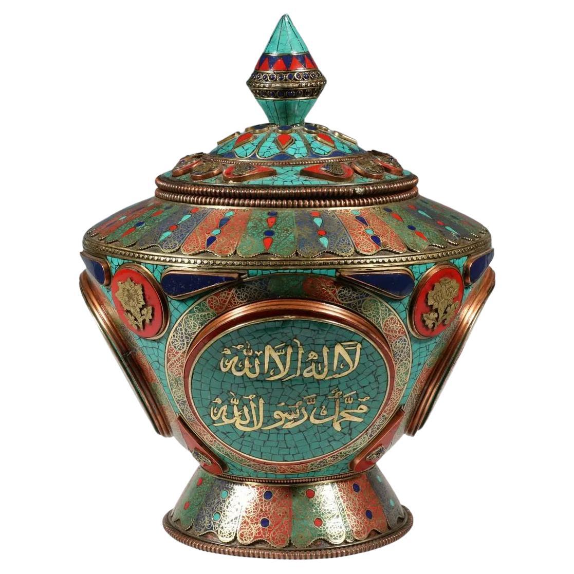 Our very unusual and large enameled metal jar or bowl with lid dates from the early twentieth century and features inlaid turquoise, lapis and carnelian stones, four cartouches with Islamic poetic calligraphy, and red, green and blue enamel in the