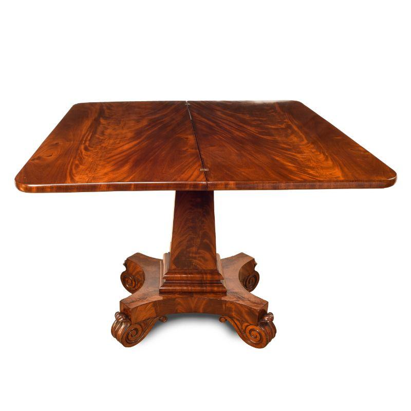American cubus mahogany flip top game table. The top rests on a truncated oblique pedestal with carved collar, joining a convex cut rectangular plinth on carved scroll feet fitted with recessed castors. Cubus mahogany solids and matched veneer