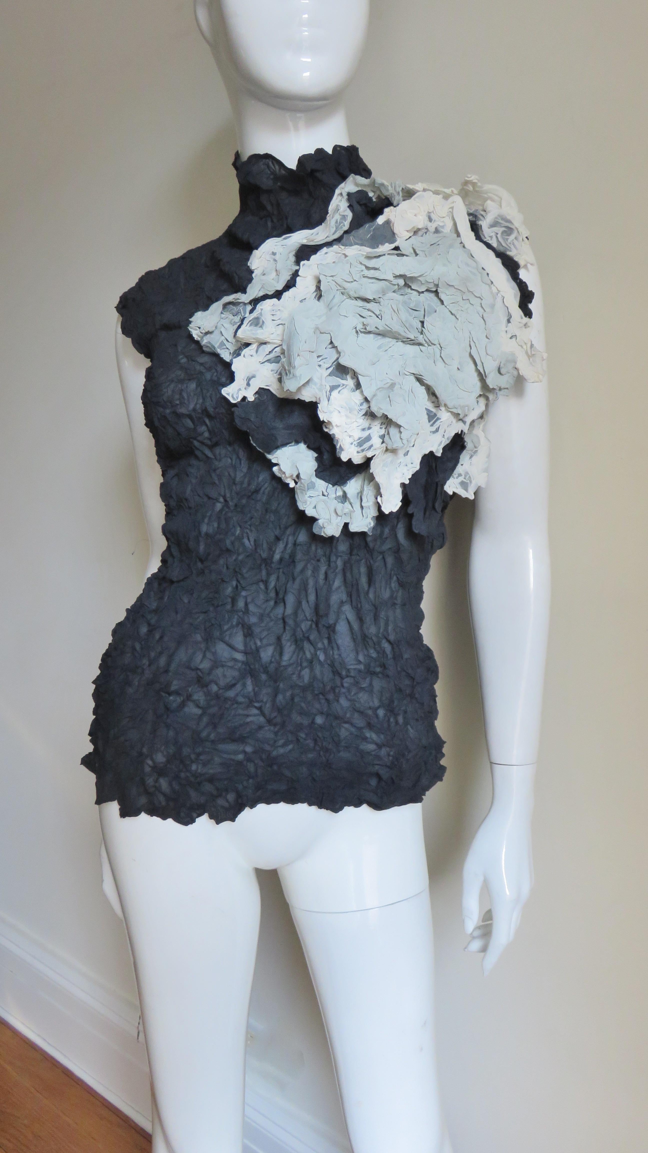 A fabulous black and grey top from Issey Miyake.  It is sleeveless with a stand up collar and semi fitted body. Off center at one shoulder is an elaborate, abstract pale taupe and grey layered flower applique.  It is very flexible in fit and slips