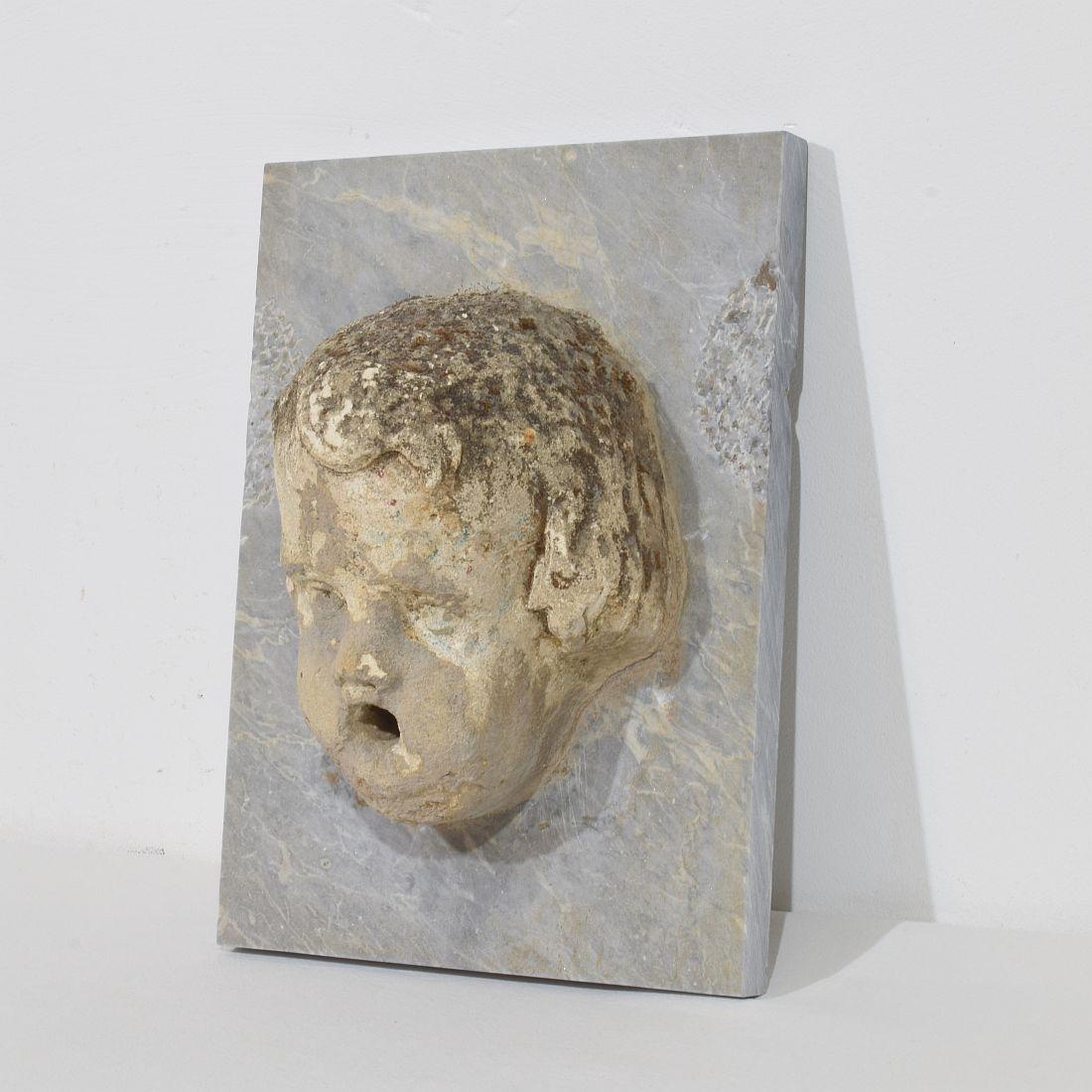 Spettacolare e unica testa di fontana di cherubino in pietra scolpita su lastra di marmo di data successiva.
Italia, 1650-1750 circa
Intemperie, piccole perdite
H. H. 39cm  L:28,5 cm P:17 cm 