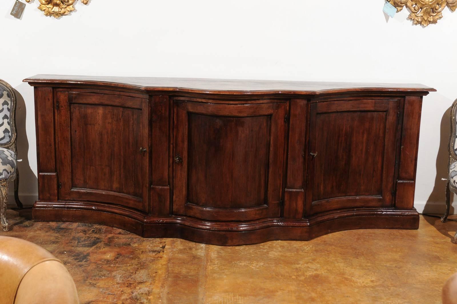 An Italian serpentine walnut three-door long buffet from the early 19th century. This Italian walnut buffet features a planked serpentine top with rounded edges, sitting above a façade made of three doors, discreetly adorned with recessed panels and