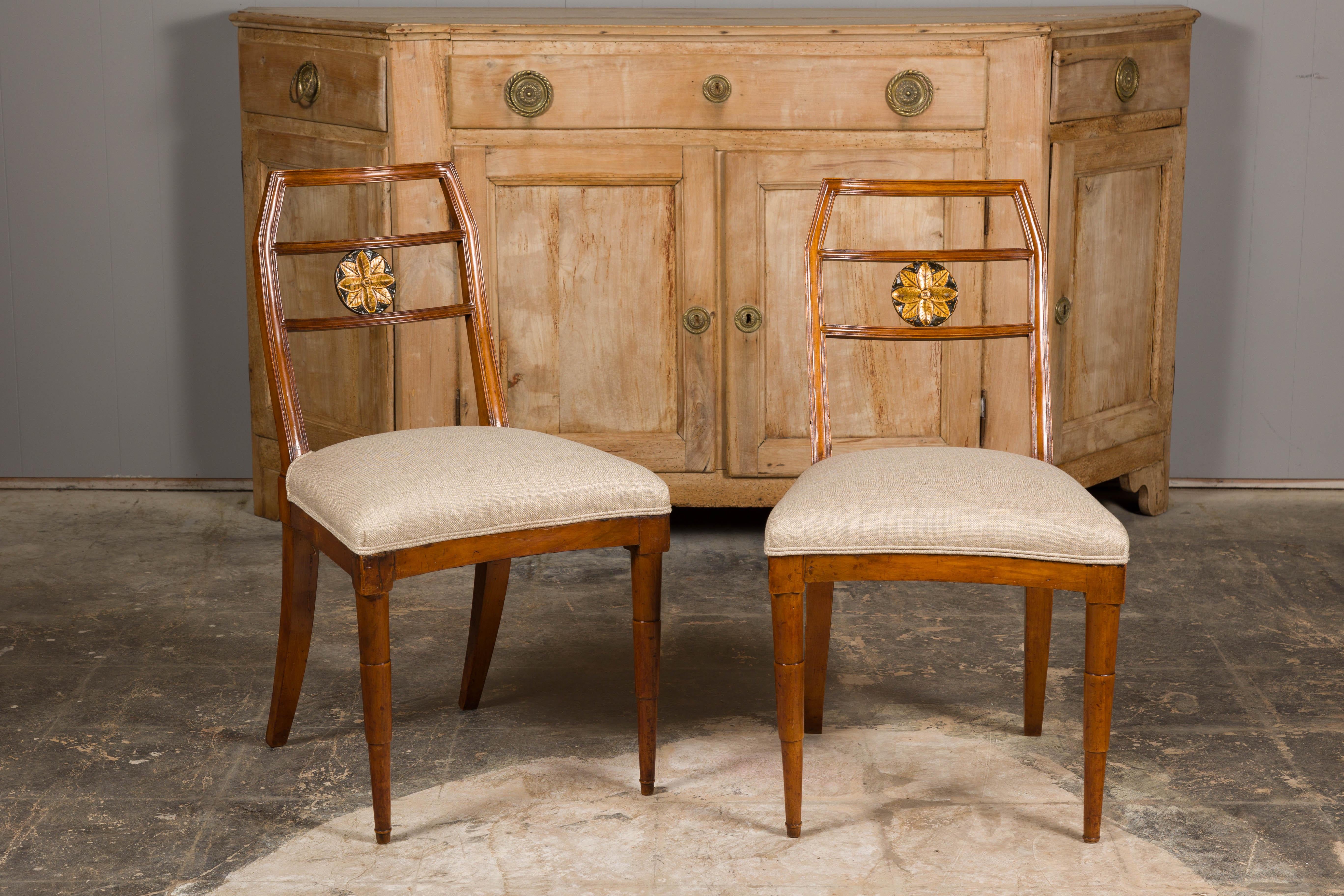 A pair of Italian walnut side chairs from circa 1800 with carved and gilt floral medallion adorning each back, custom linen upholstery and tapering legs in the front. This pair of Italian walnut side chairs, dating back to circa 1800 is a