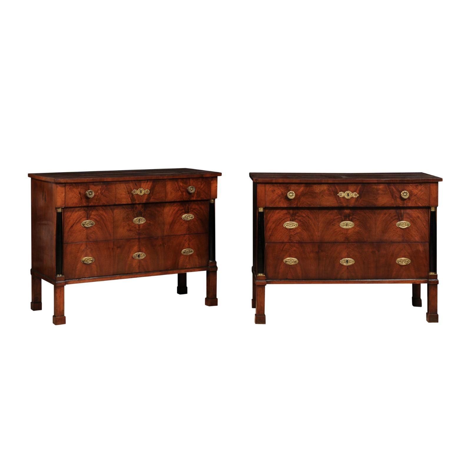A pair of Italian Empire period walnut and mahogany commodes from circa 1820 with three drawers, bookmatched veneer, ebonized columns and bronze hardware. Imbue your home with the rich patina of history with this exquisite pair of Italian Empire