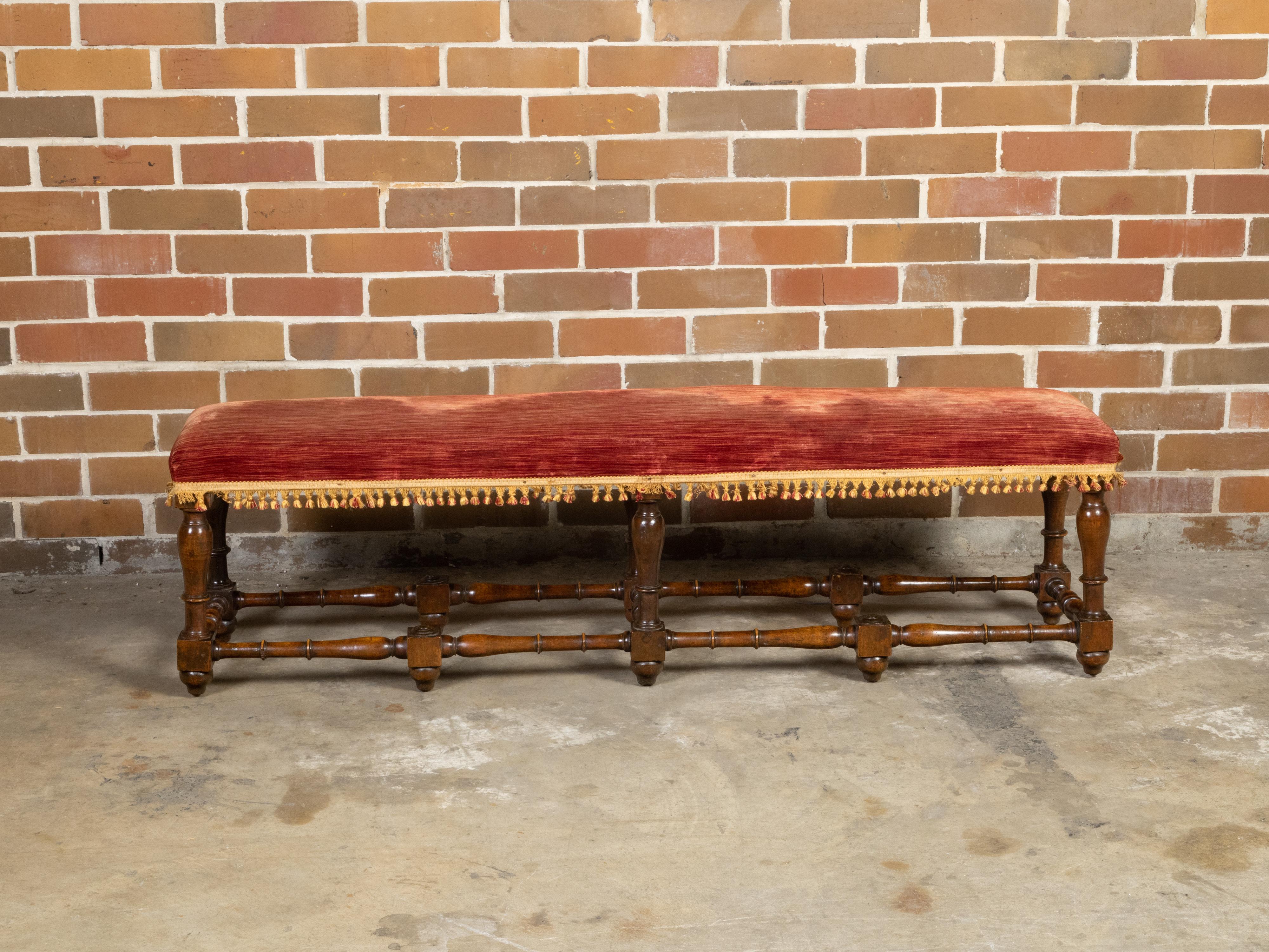An Italian walnut bench from the early 19th century, with red fabric, petite tassels, turned legs and stretchers. Created in Italy during the first quarter of the 19th century, this walnut bench features a long rectangular seat covered with a red