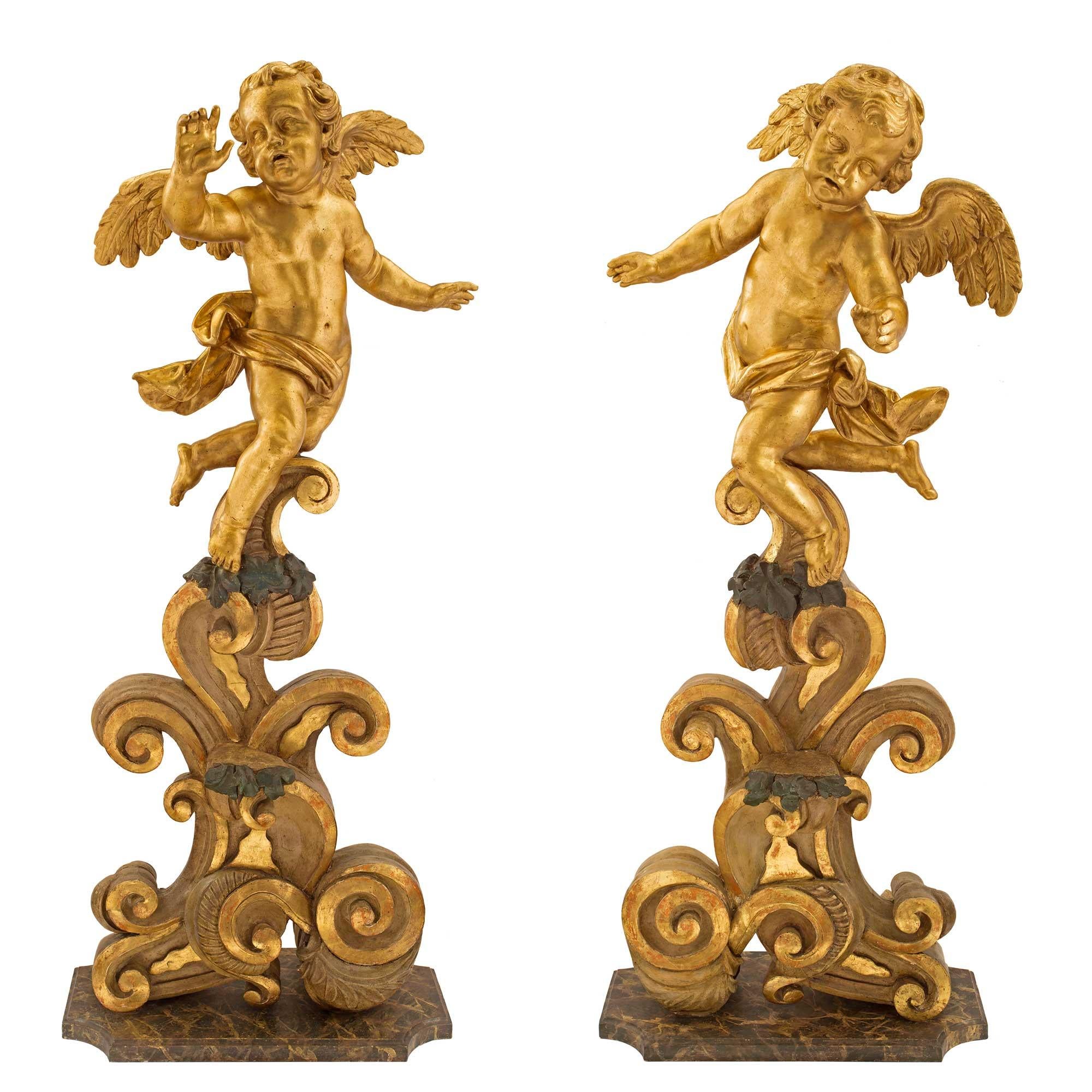 A sensational and extremely decorative true pair of Italian 18th century Baroque giltwood, polychrome and faux painted marble cherub statues. Each impressive and large scale statue is raised by a beautiful rectangular base with concave corners and a