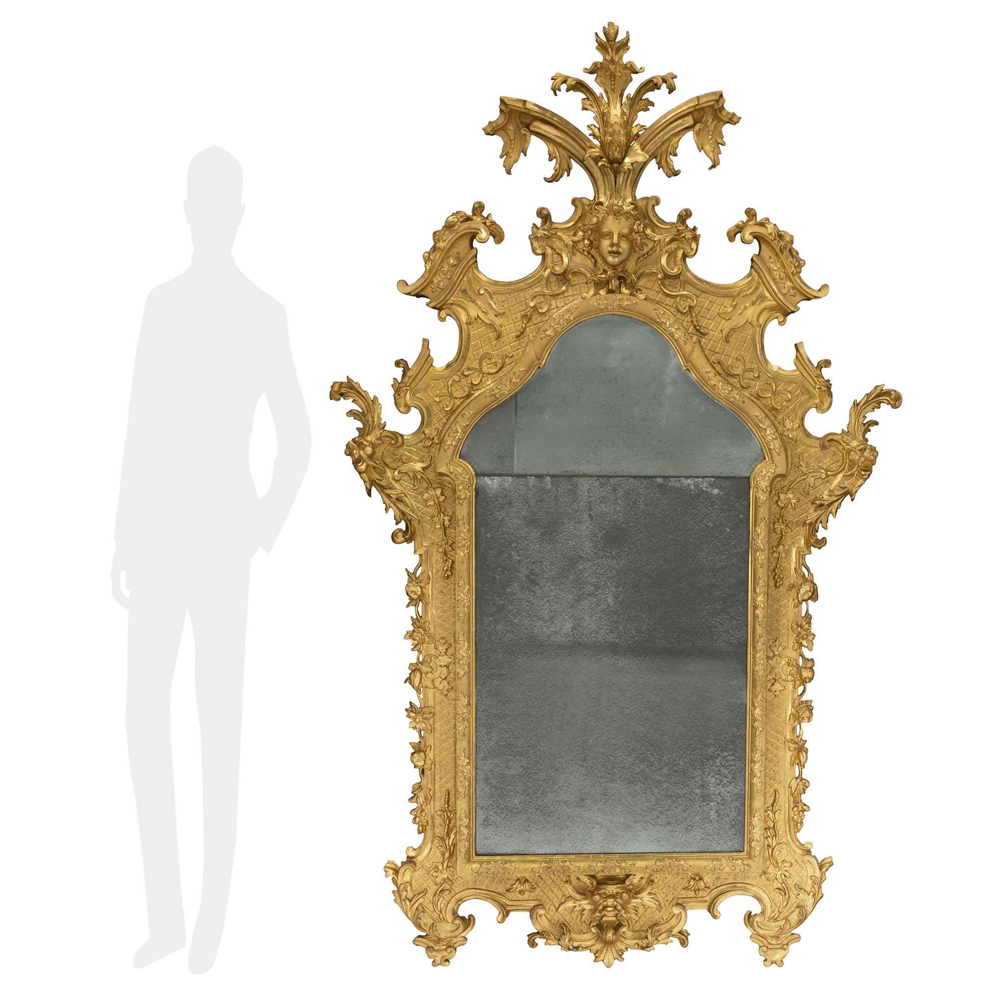 A stunning and large scale Italian 18th century Baroque period giltwood mirror. The original mirror plates are framed within a foliate and hammered designed mottled border. At the base are foliate feet with rich scrolled patterns which center a