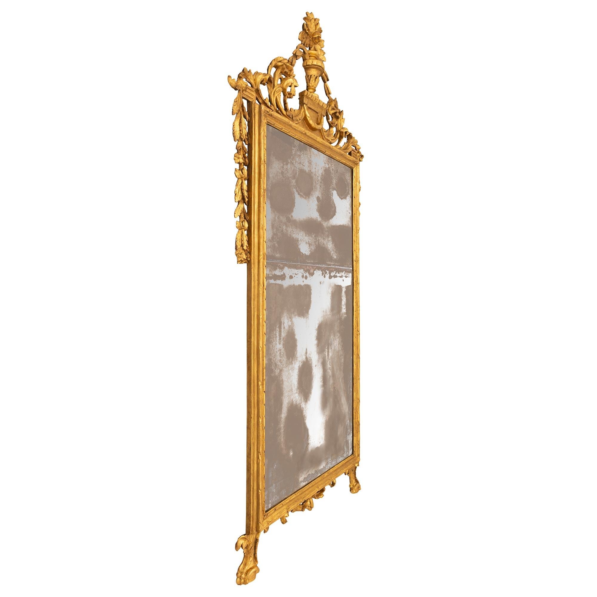 An elegant and large scale Italian 18th century Louis XVI st. giltwood mirror. The mirror from the Genovese region is raised by two supporting legs flanking the central reserve of scrolled acanthus leaves. The rectangular mottled giltwood frame is