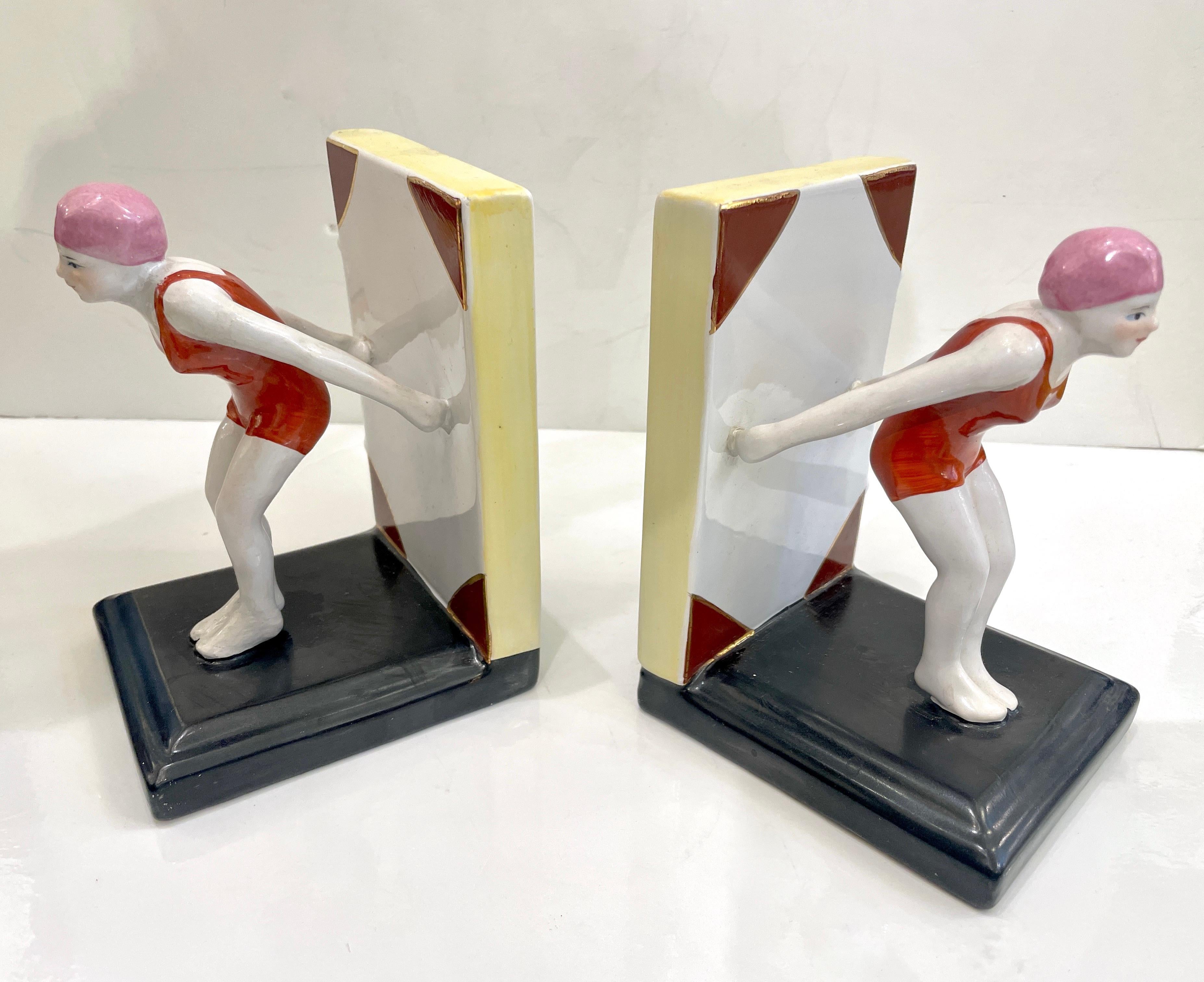 Un'impressionante coppia di reggilibri in ceramica Art of Vintage che raffigurano eleganti tuffatrici, ognuna in posizione dinamica. Questi fermalibri scultorei raffigurano donne in costume da bagno rosso e cuffia rosa, dipinte a mano con dettagli