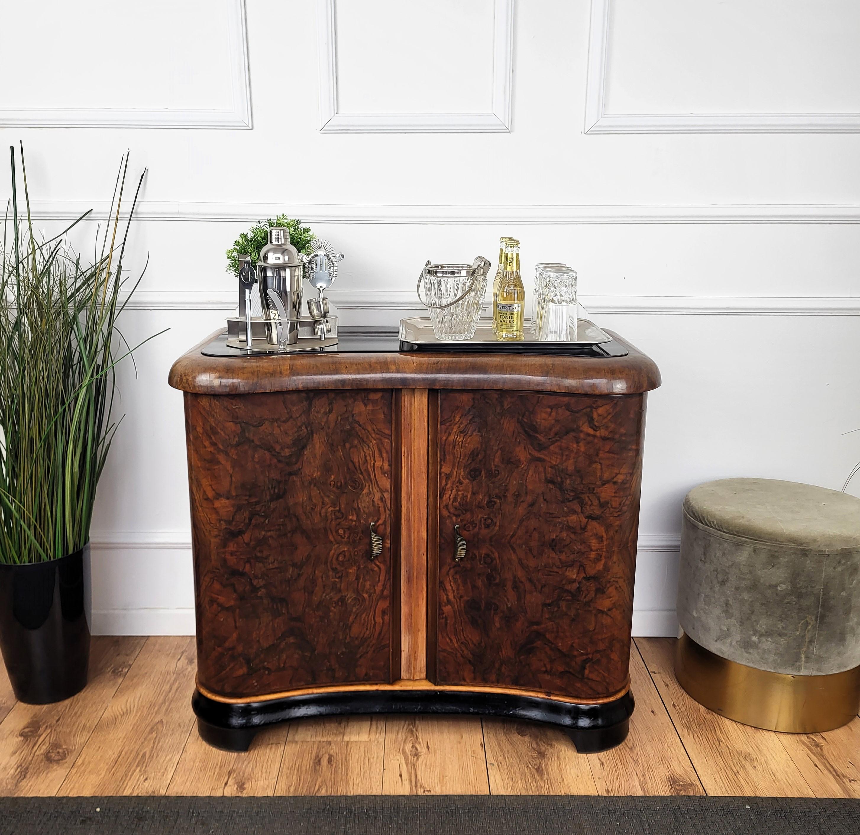 Very elegant Italian Art Deco Mid-Century Modern drinks dry bar cabinet with beautiful veneer walnut briar burl wood, two doors and great central wooden decor craftmanship, brass handles and framed black glass top. When opened, on the right side an