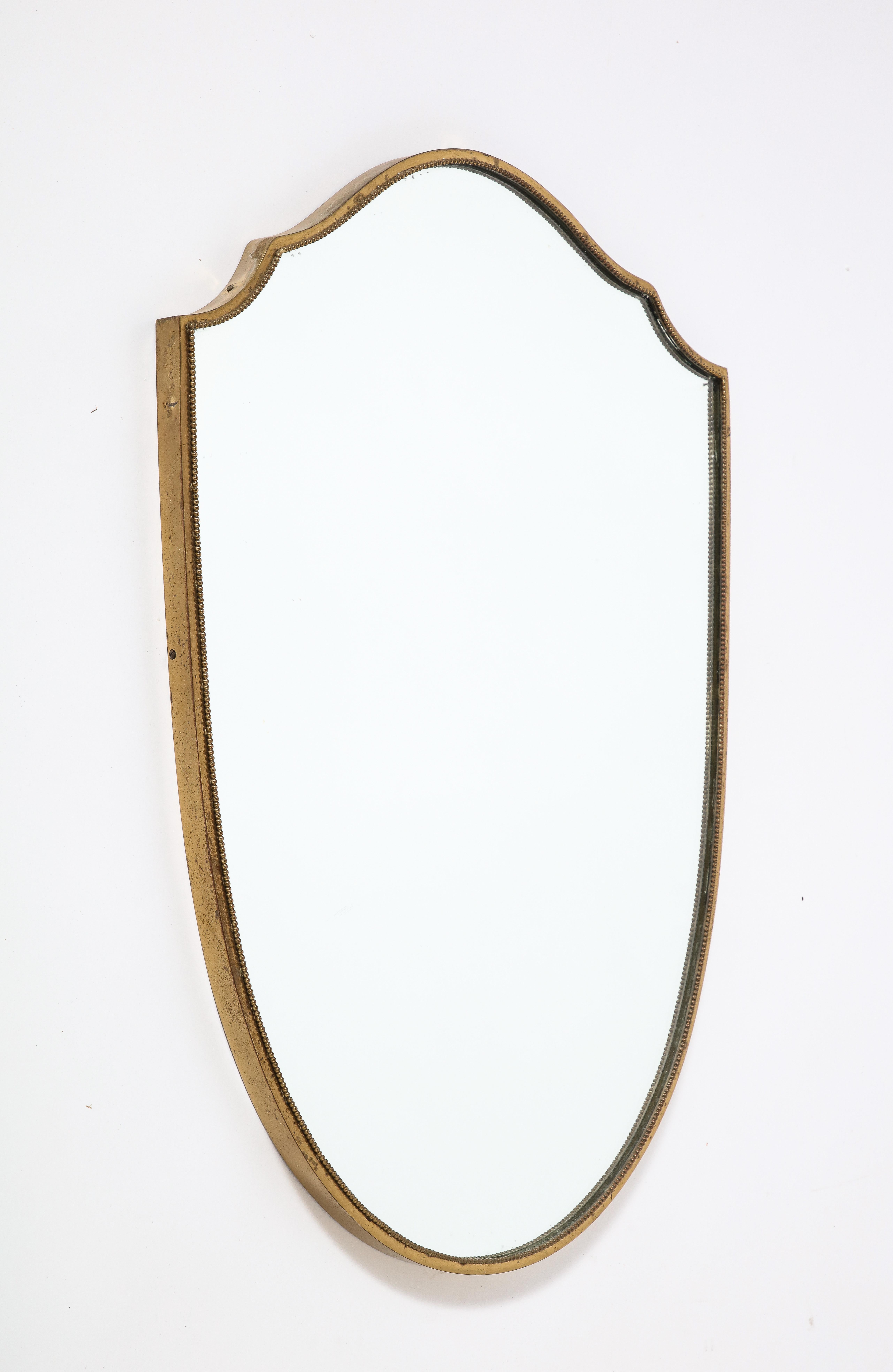 An Italian 1940's brass shield shaped mirror with beaded trim; the brass with a warm patina, mirror glass plate original. 
Italy, circa 1940 
Size: 31 1/2