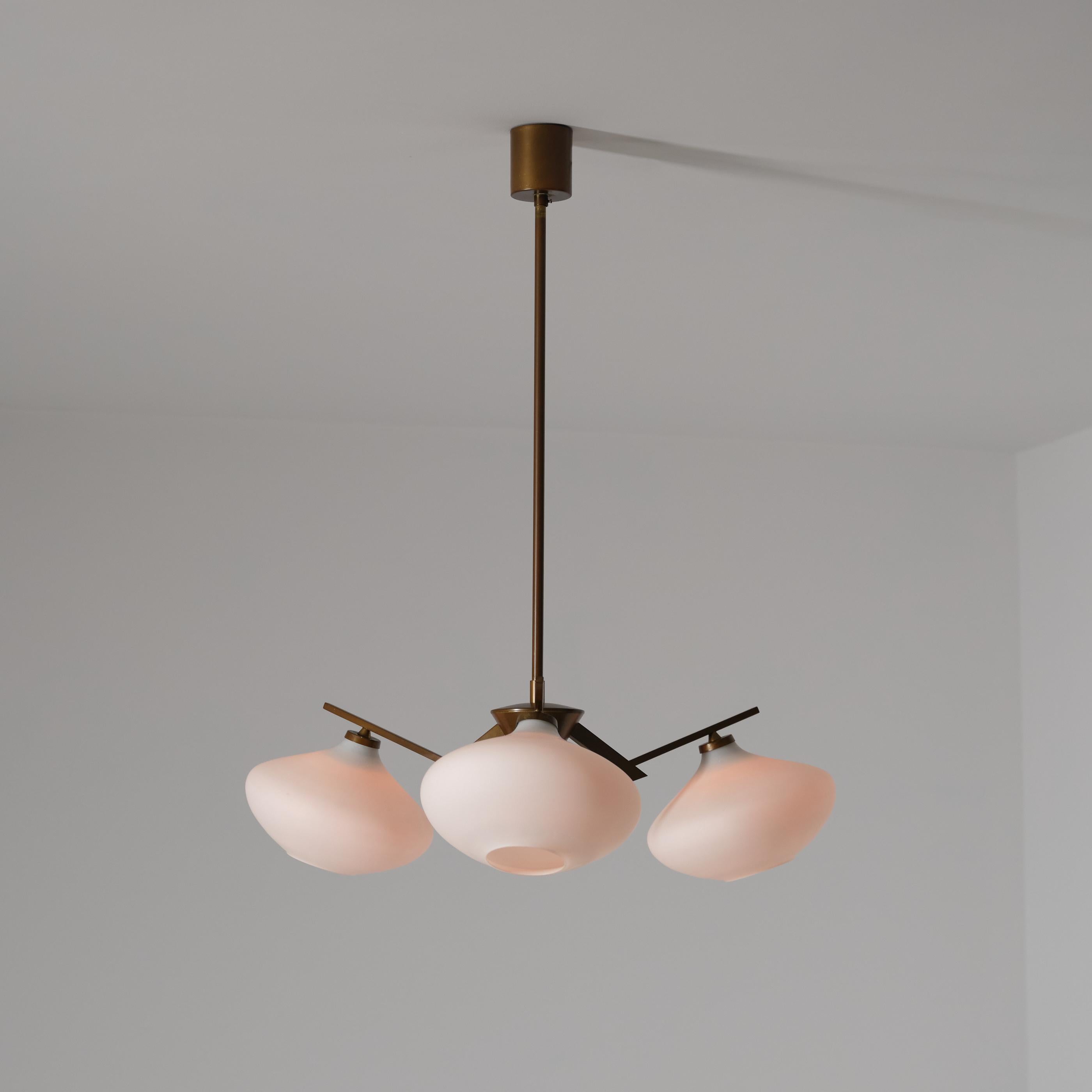 Elegant Italian chandelier from the 1950s, featuring a sculptural brass structure with a beautiful, even patina and three hand-blown opaline glass shades. The arms radiate from a central hub in a refined geometric composition, while the softly