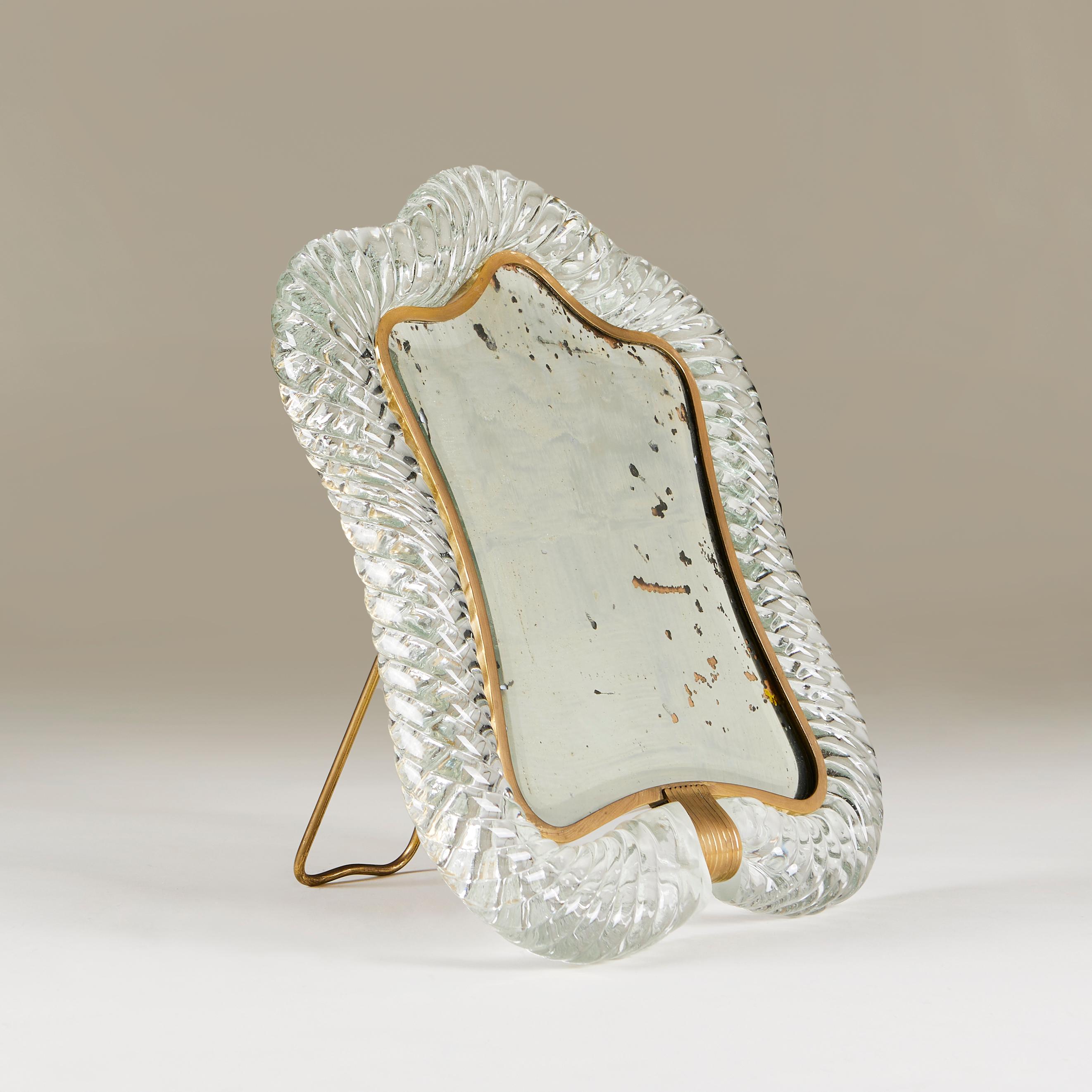 Geformter Tischspiegel von Barovier & Toso mit Rahmen aus gedrehtem Murano-Glas (Torchon). Der Rahmen ist von einem dünnen Messingfilet und einer Messingschließe umgeben. Rückenstrebe aus Messing mit signierter Herstellermarke. Der Spiegel, der