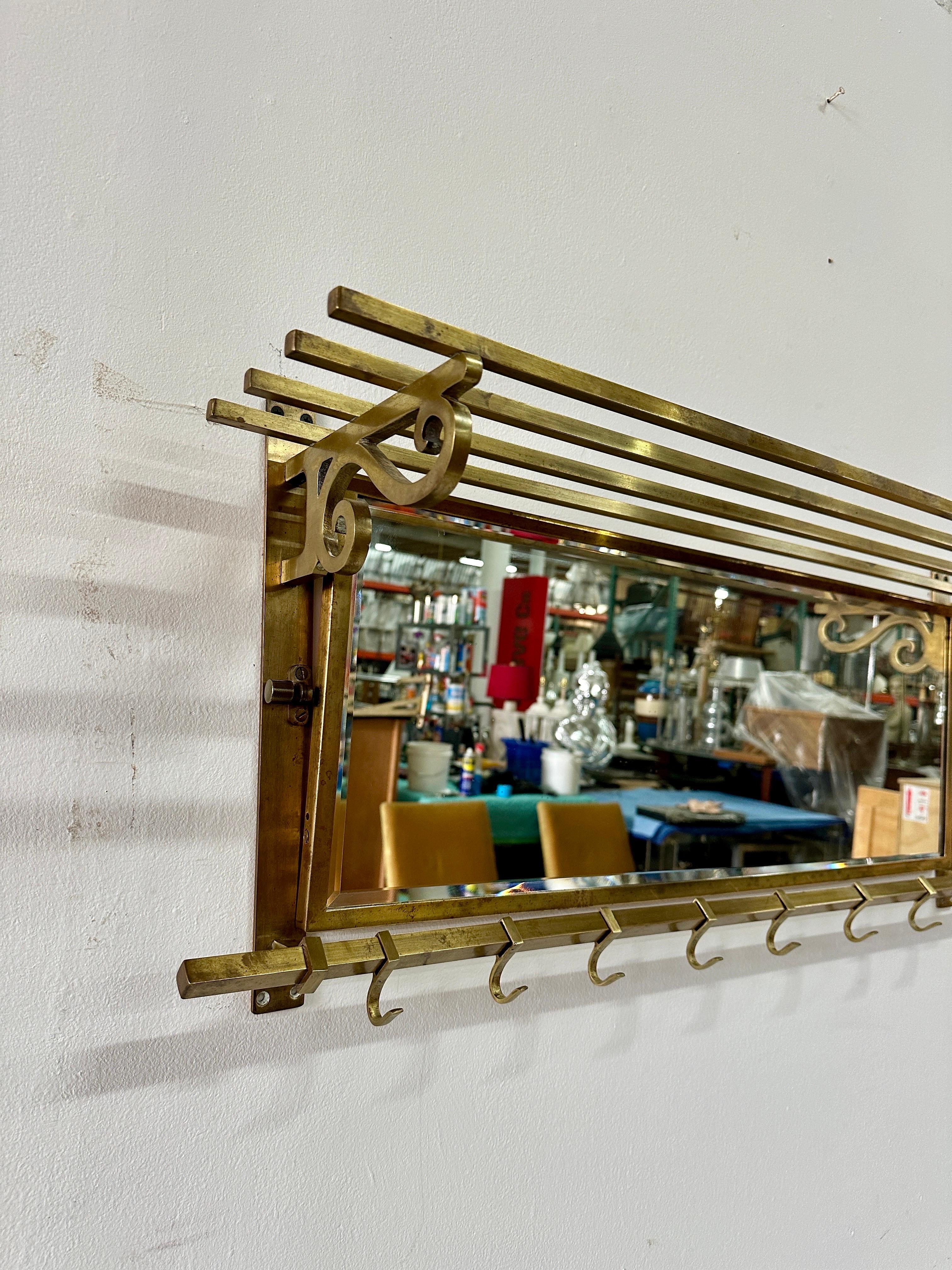 Questo grazioso appendiabiti vintage italiano montato a parete in ottone e specchio è dotato di ganci in ottone che scorrono lungo il ripiano inferiore per appendere gli oggetti più grandi.  Lo specchio in ottone è montato su un supporto girevole e