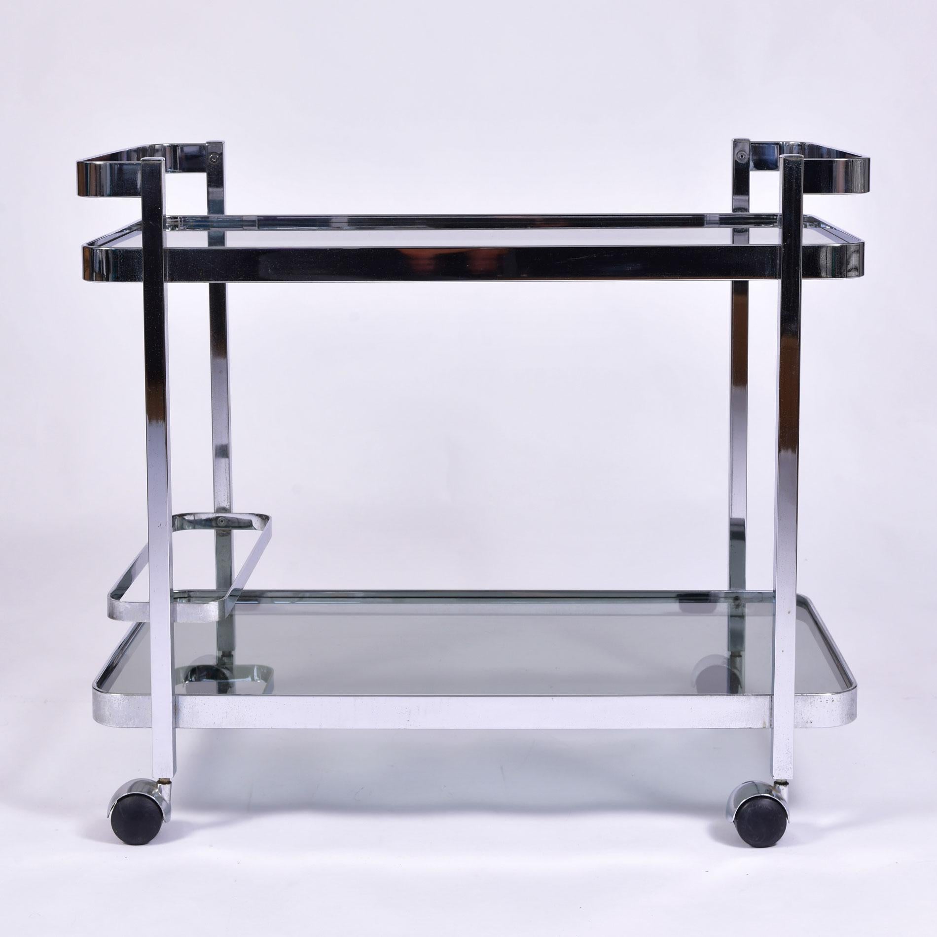 Chic chrome drinks trolley with two smokey glass shelves. Shelves are surrounded by simple curved chrome frame extending on the top shelf to create a handle at both ends. Bottle holder holds up to three bottles. Sits on chrome castor wheels for ease