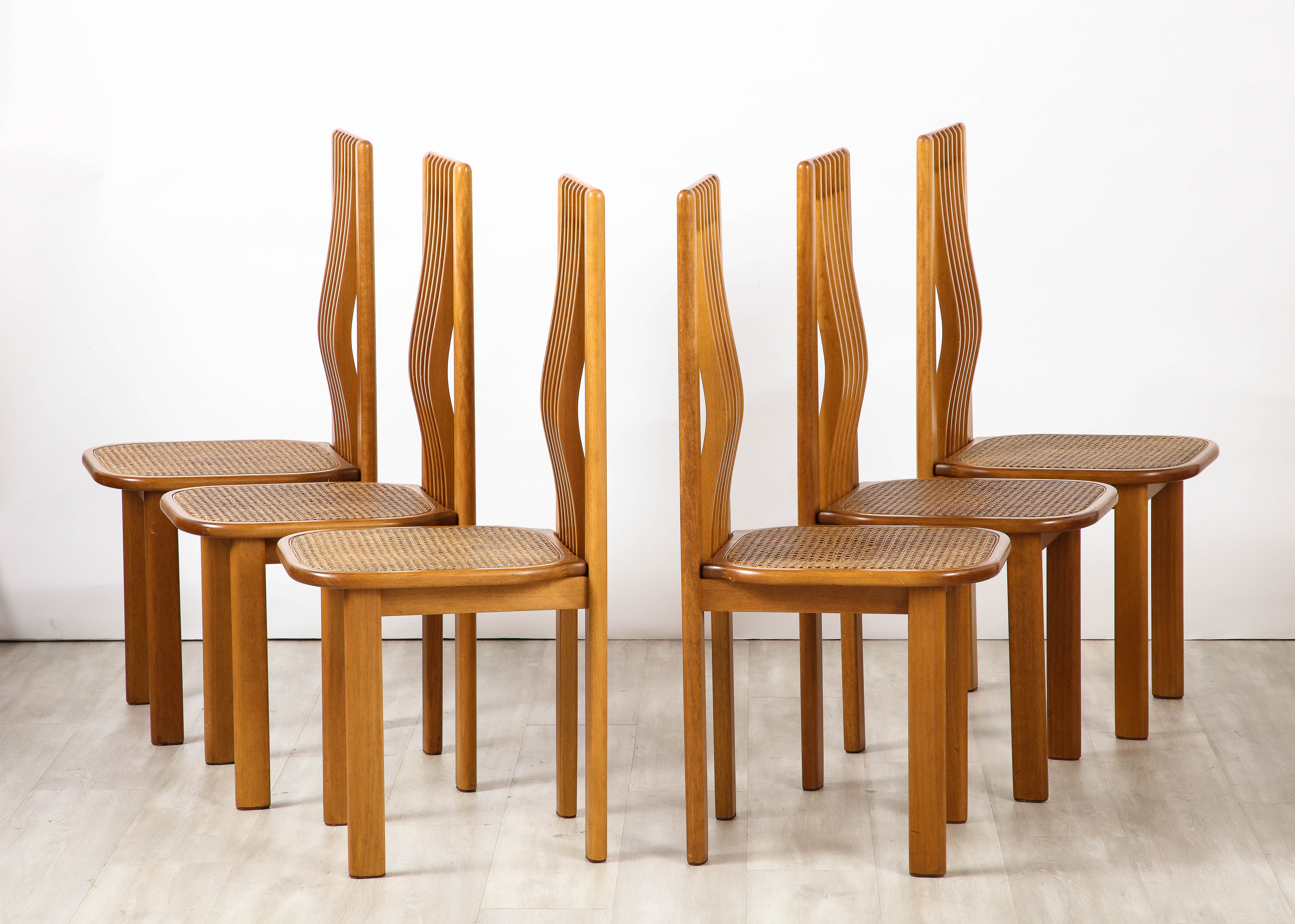 An Italian 1970's set of six dining chairs in maple with caned seats, the high backs are slatted with the mid-section indented forward, molding to your back. The cane seats extend slightly over the angled legs.   They are highly sculptural, sleek