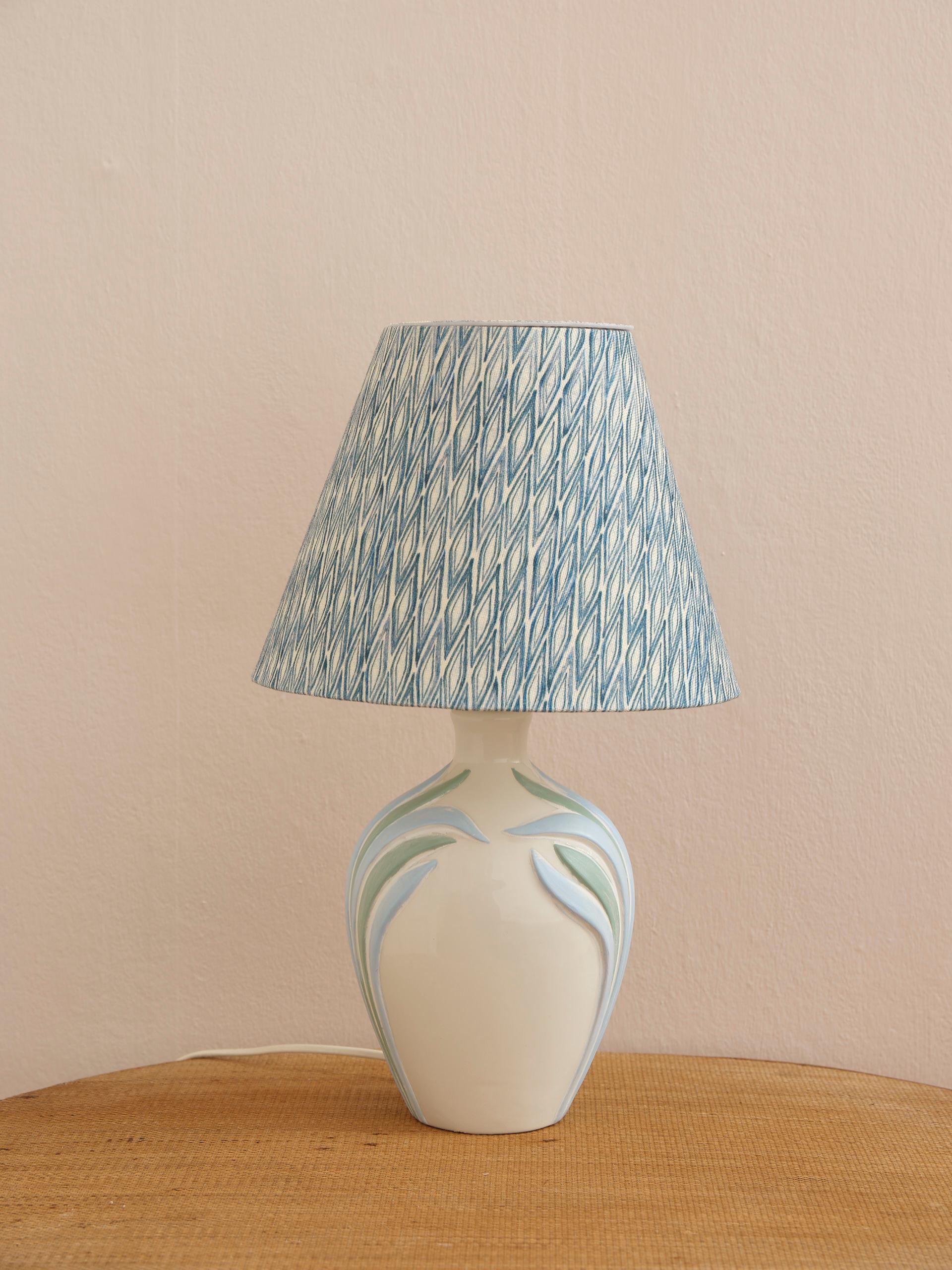 Esta preciosa lámpara de mesa italiana de porcelana mate está decorada con hojas de porcelana azul pastel y verde que la convierten en una pieza luminosa típica de los años 80 y atemporal a la vez. Aquí, montada con una pantalla de lámpara hecha a