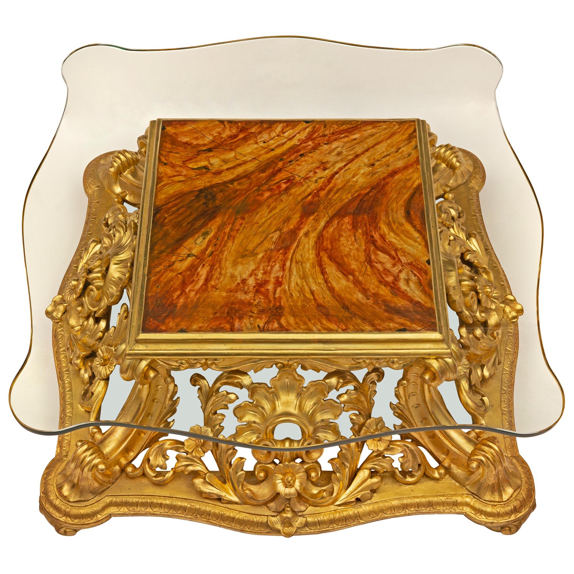 A stunning and very unique Italian 19th century Baroque st. giltwood, faux painted marble, and glass coffee table. The table is raised by an exceptional and most decorative scalloped shaped giltwood base with ball feet and exquisite richly carved