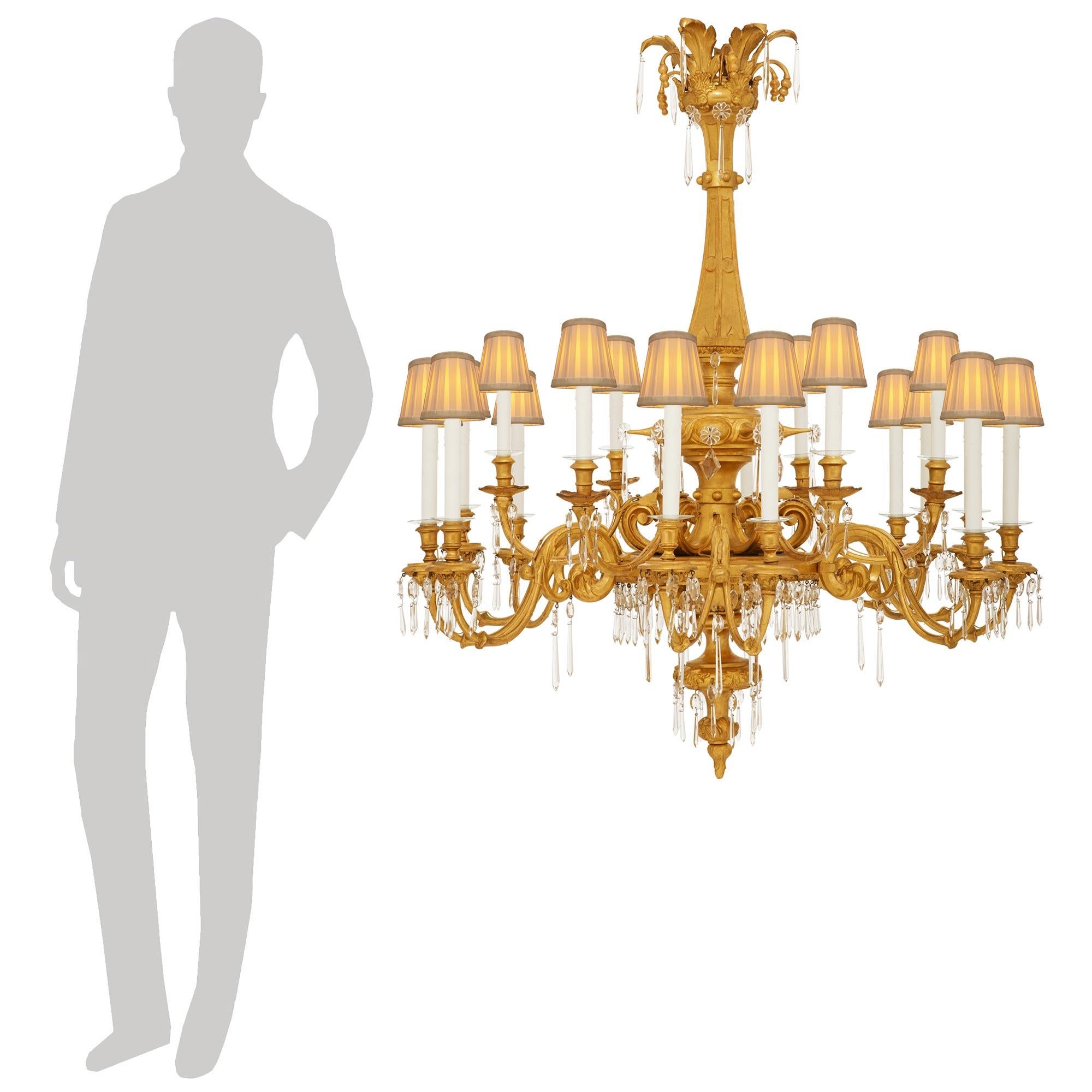 Un lustre impressionnant et très décoratif en bois doré et cristal italien du 19ème siècle. Ce magnifique lustre à dix-huit bras de la région de Lombardie est centré par un épi de faîtage inversé décoré de grandes palmettes et de motifs feuillus.