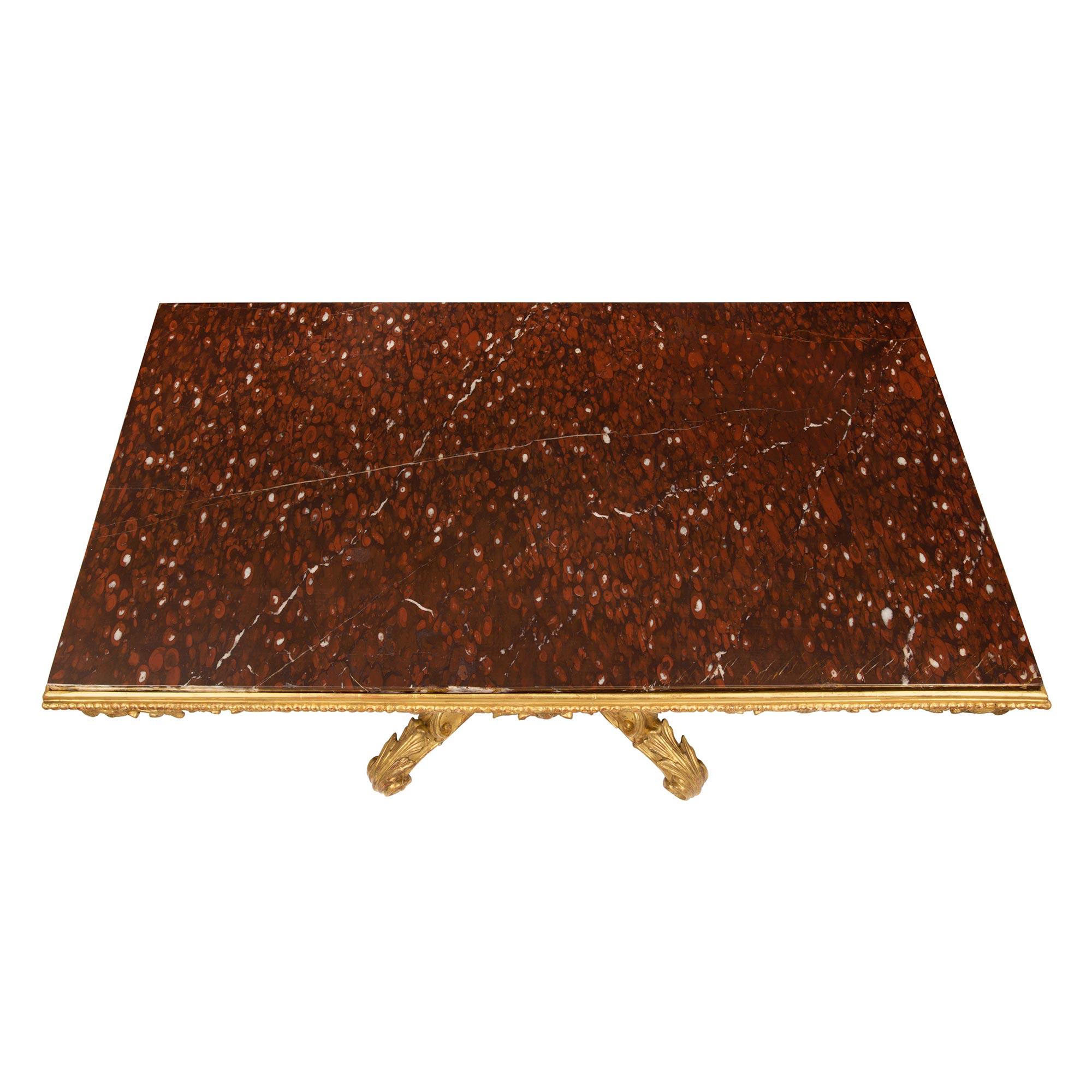 Impresionante mesa de centro rectangular italiana de principios del siglo XIX de madera dorada y mármol Rouge Griotte. La mesa se eleva sobre un pedestal de cuatro patas con roleos, opulentamente decorado con tallas de máscaras y grandes hojas de