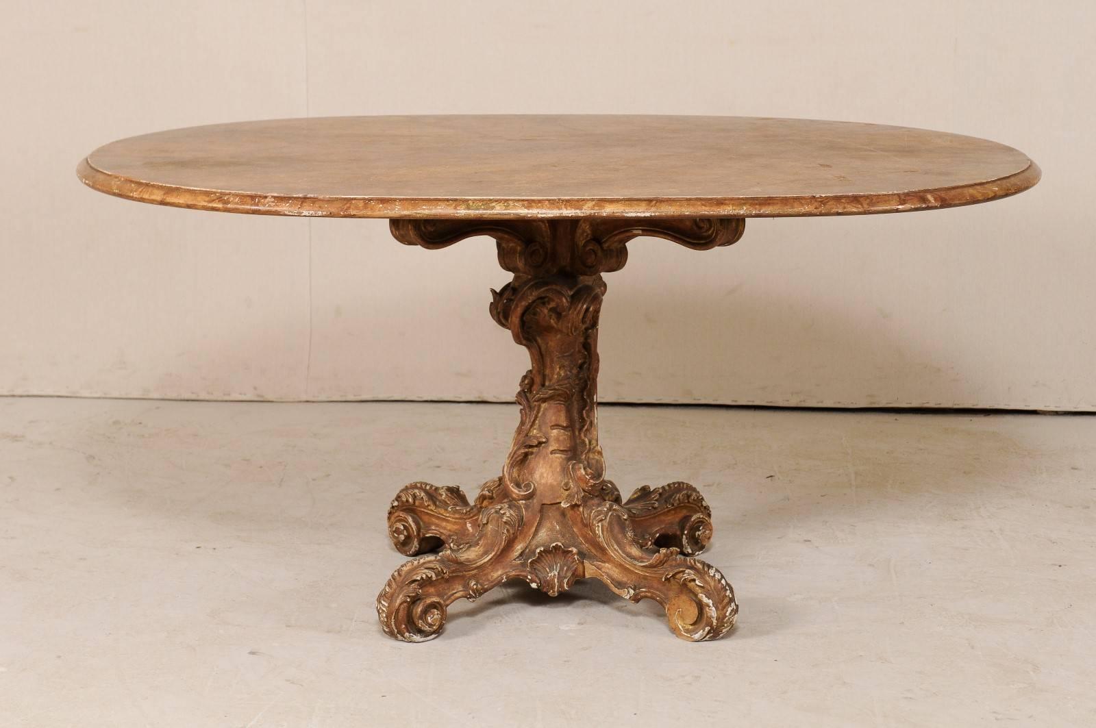 An Italian 19th century oval pedestal table with ornately carved wood base. This antique table from Italy features an oval-shaped top with a faux marble finish, resting atop a curvy and naturalistic style pedestal base carved with acanthus leaves