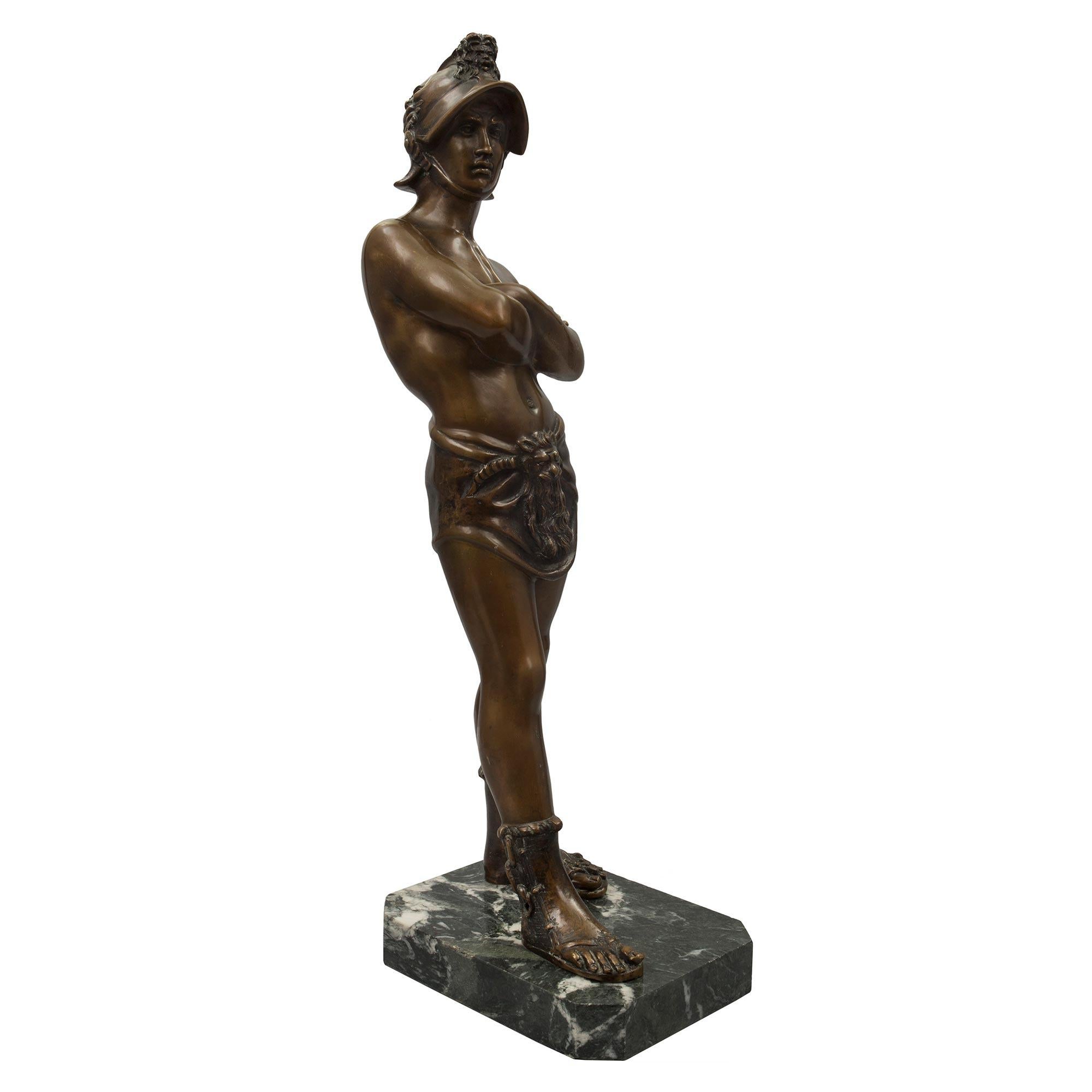 Belle statue italienne en bronze patiné du XIXe siècle représentant un jeune gladiateur. La statue est surélevée par un socle rectangulaire en marbre Verde Antico. Le gladiateur se tient fièrement debout, les bras croisés, et porte des sandales à
