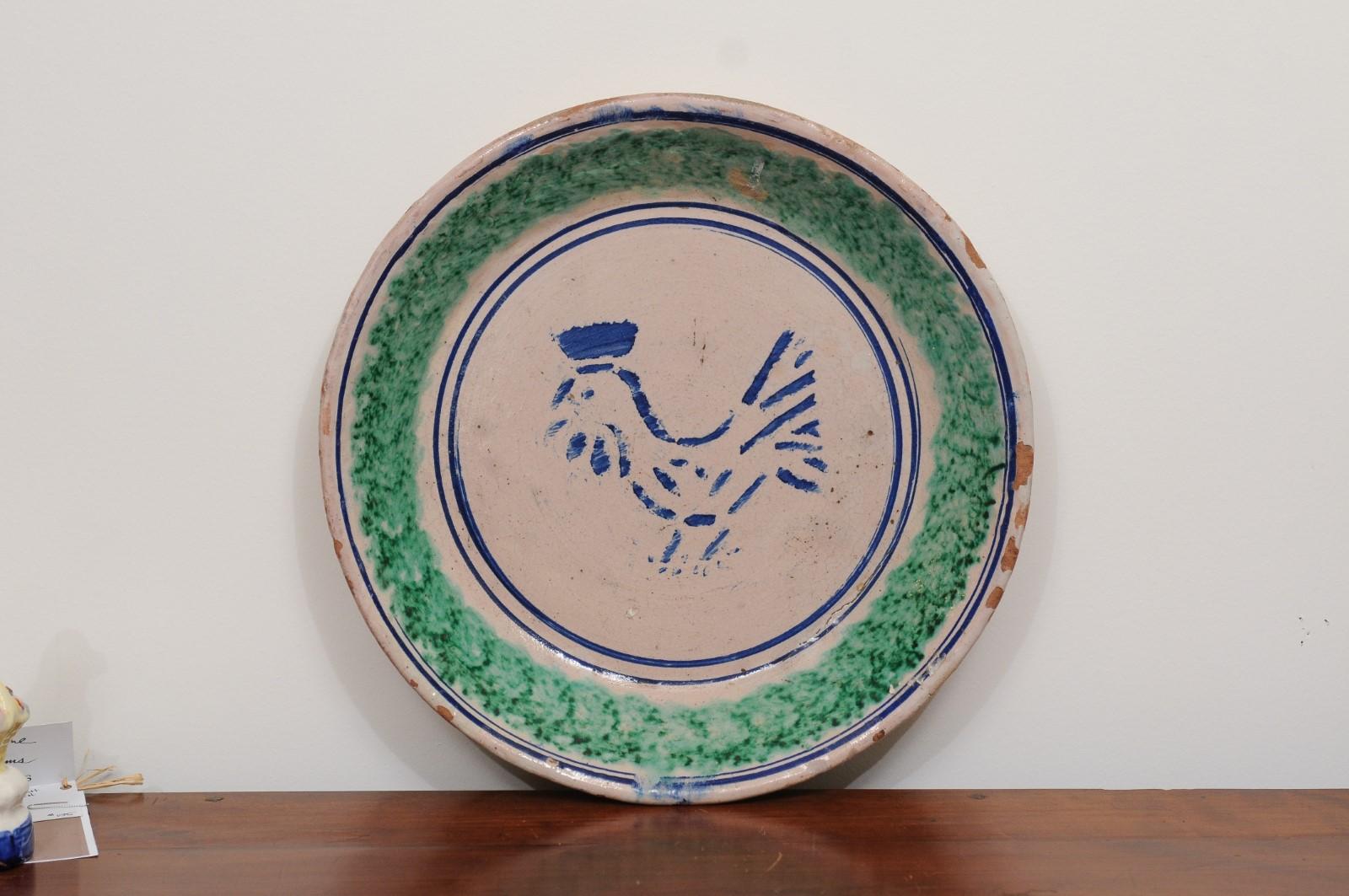 An Italian pottery plate from the 19th century, with stylized rooster and green accents. Created in Italy during the 19th century, this pottery plate features blue and green tones alternating with one another. A blue rooster is depicted in profile