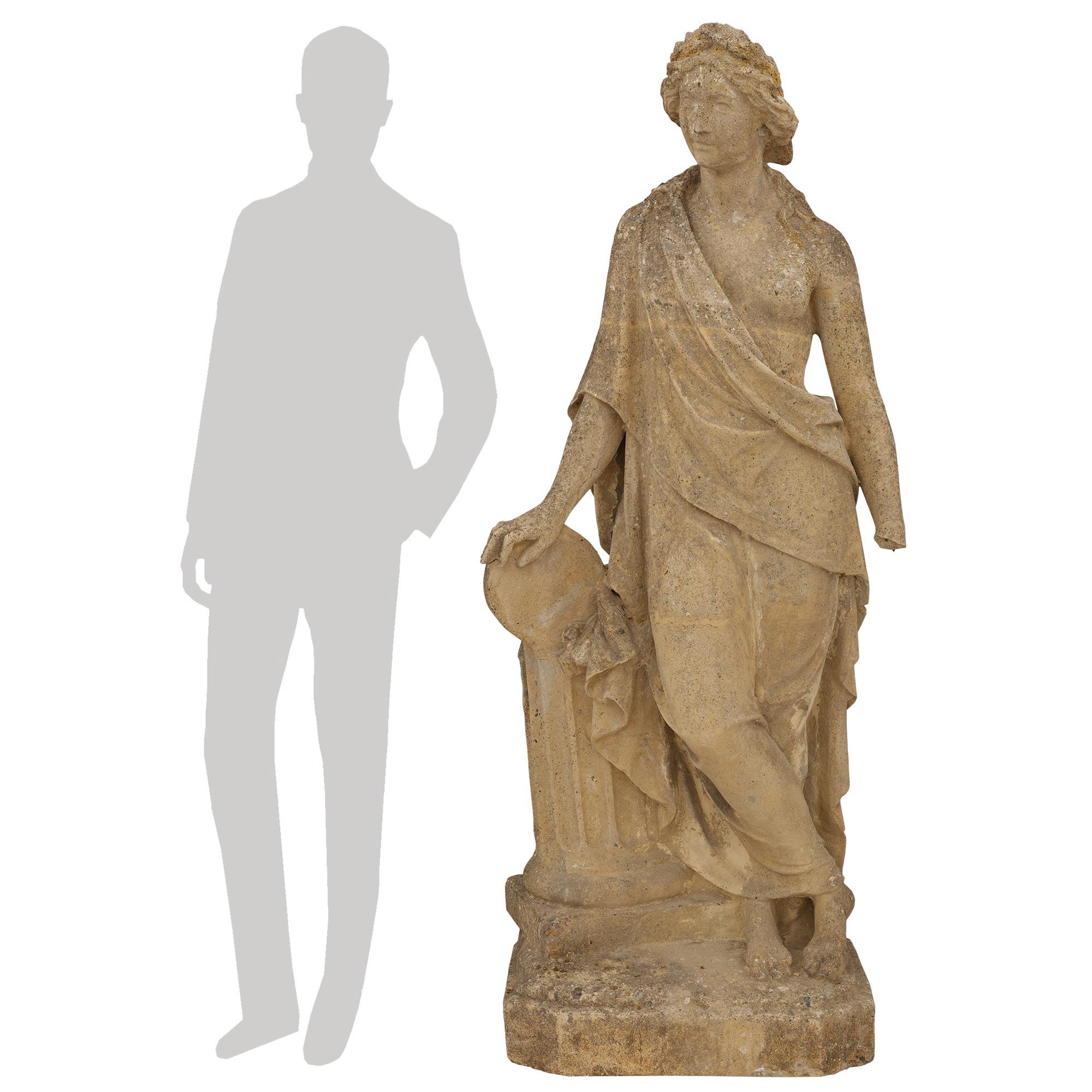 Una bella statua italiana in pietra del XIX secolo. La figura femminile classica è sollevata su una base quadrata in pietra con angoli tagliati. È appoggiata a una colonna scanalata con le gambe incrociate. La figura è adornata in abito classico con