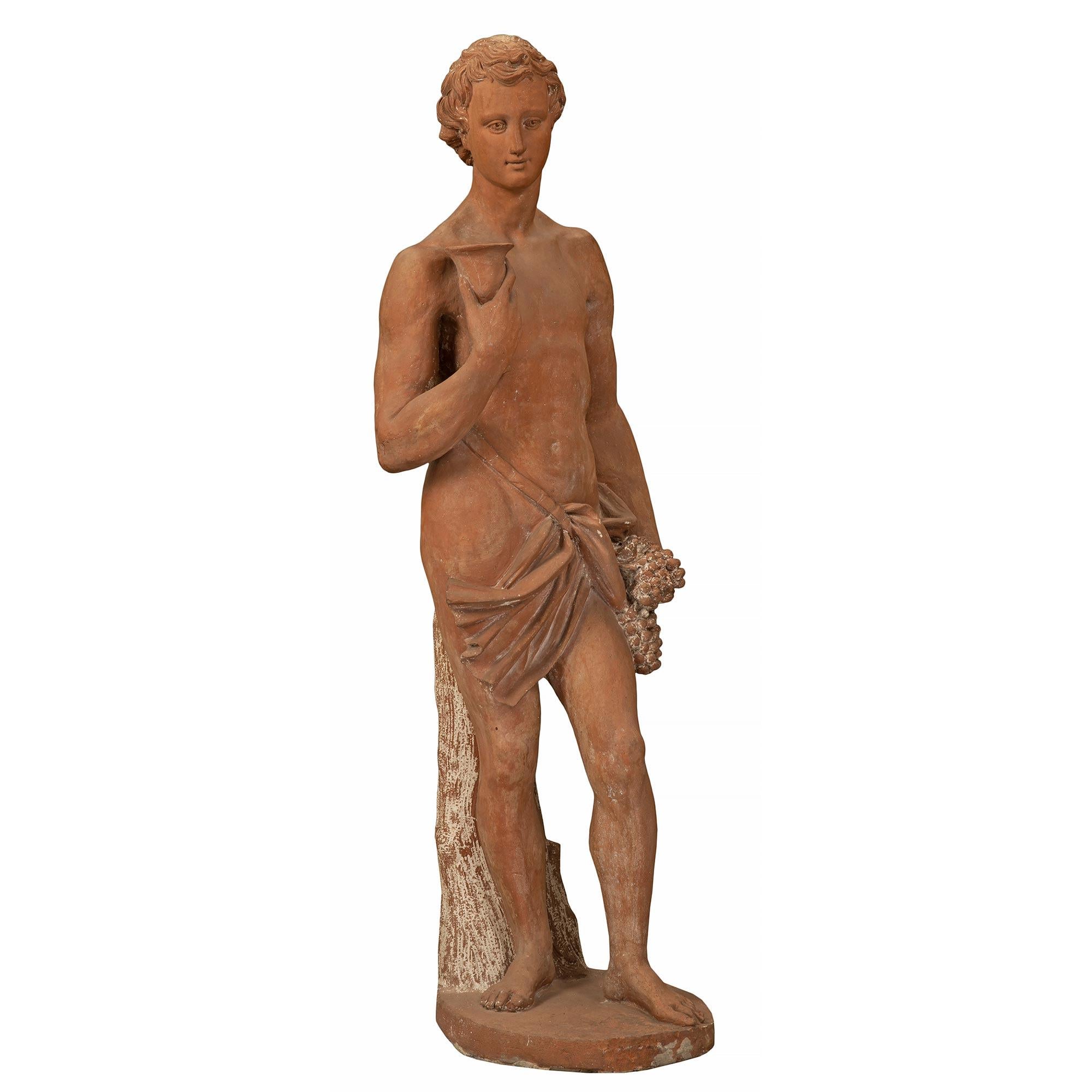 A charming Italian 19th century terra cotta statue of a young Bacchus. The statue is raised by an oblong base where the handsome young man stands in front of a tree trunk. He has a pouch at his hip that is strapped over his shoulder. Bacchus holds