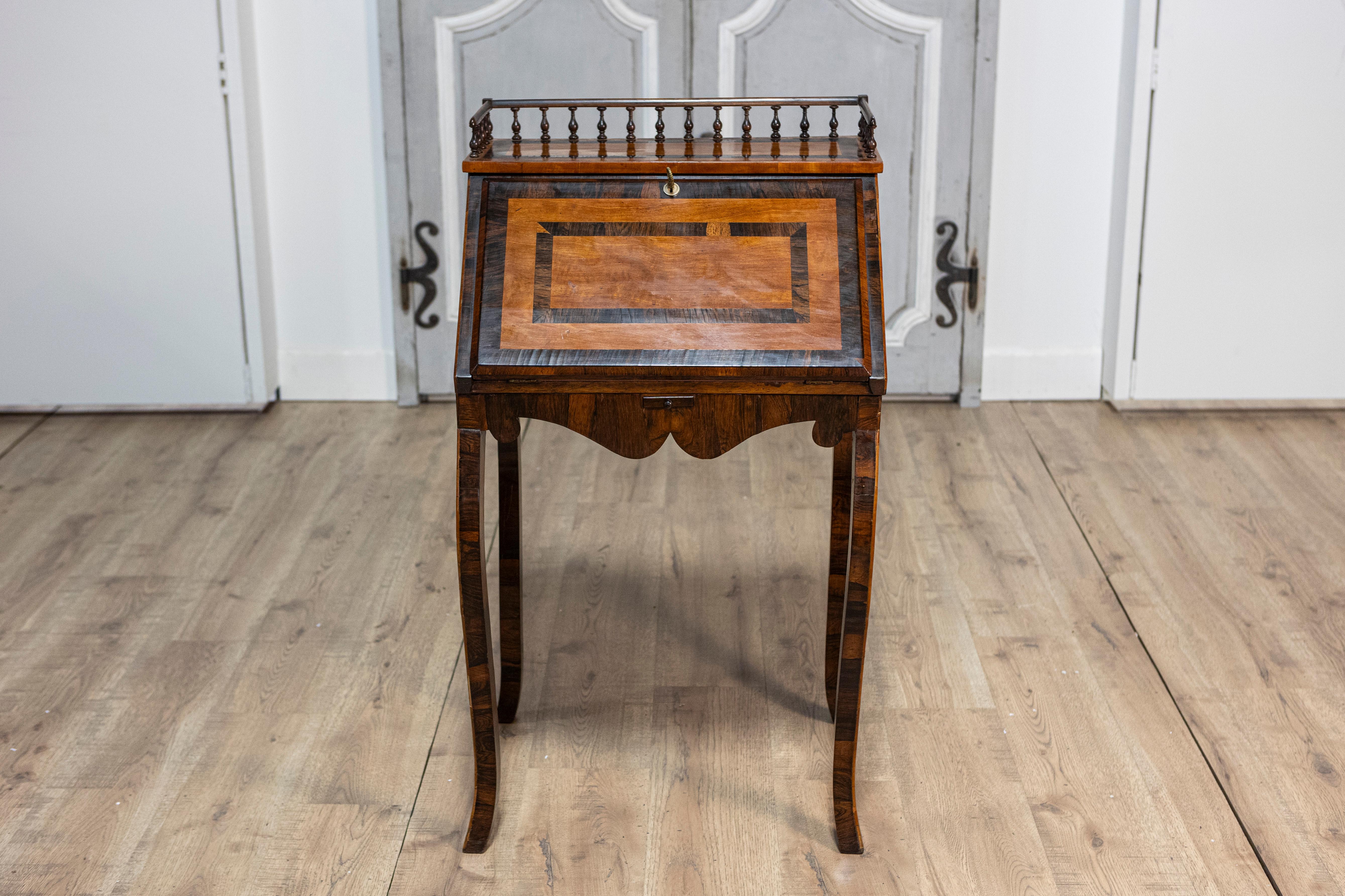 A petite Italian two-toned walnut and mahogany writing desk from the 19th century with slant front, turned three-quarter gallery and carved apron. This petite Italian two-toned walnut and mahogany writing desk from the 19th century is a charming