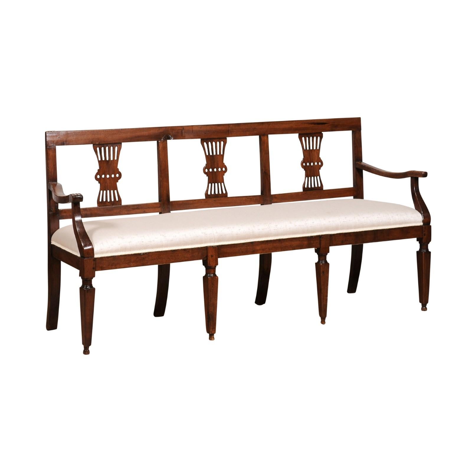 An  Italian walnut three-seater bench from the 19th century with carved splats, curving open arms, tapered legs in the front and saber ones in the back. Experience the timeless elegance of Italian craftsmanship with this captivating walnut