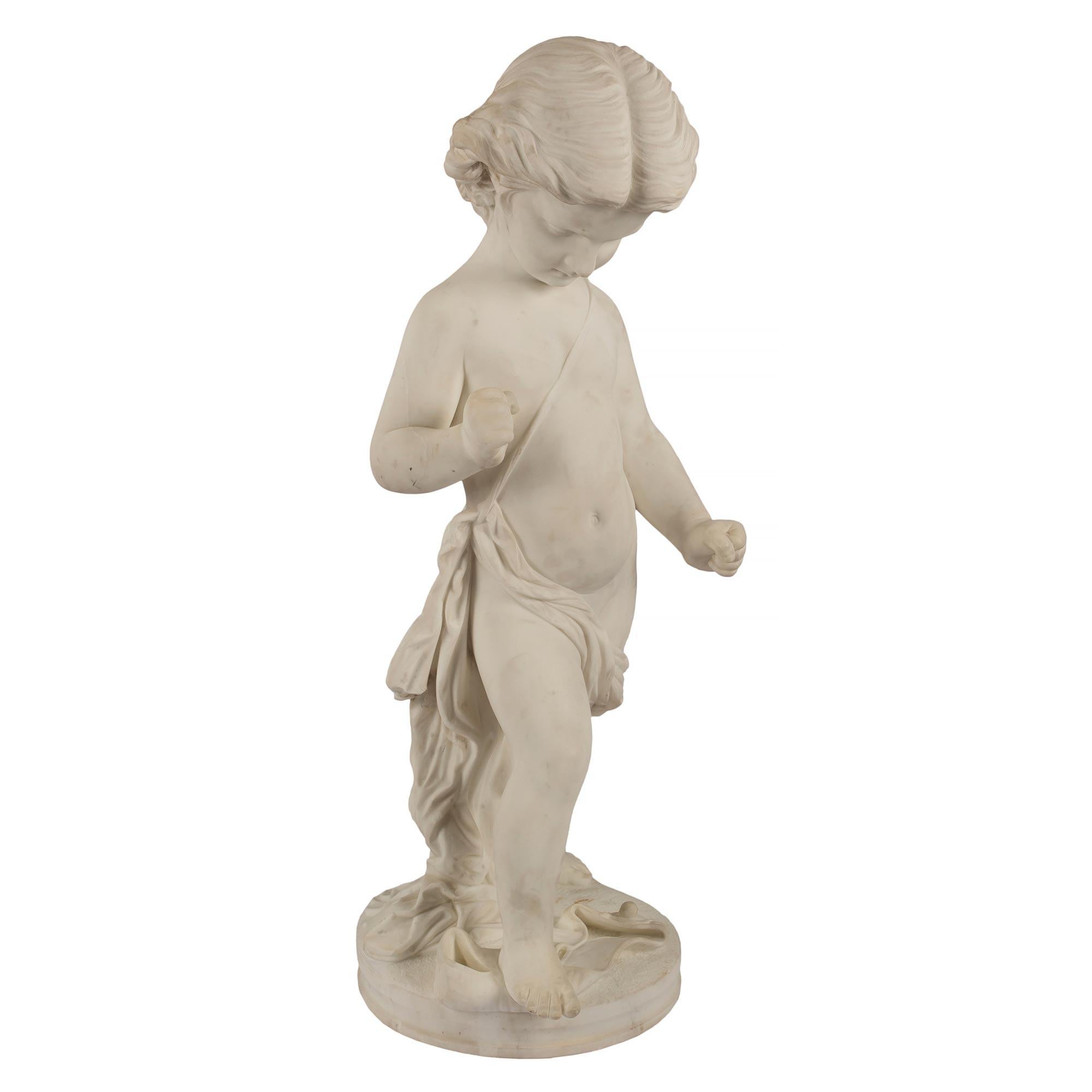 A charming Italian 19th Century white Carrara marble statue of a young boy. The boy is raised by a circular base seeming to be slightly irritated by the fact that his belt has fallen. While standing and clenching his wrists out of frustration, he is