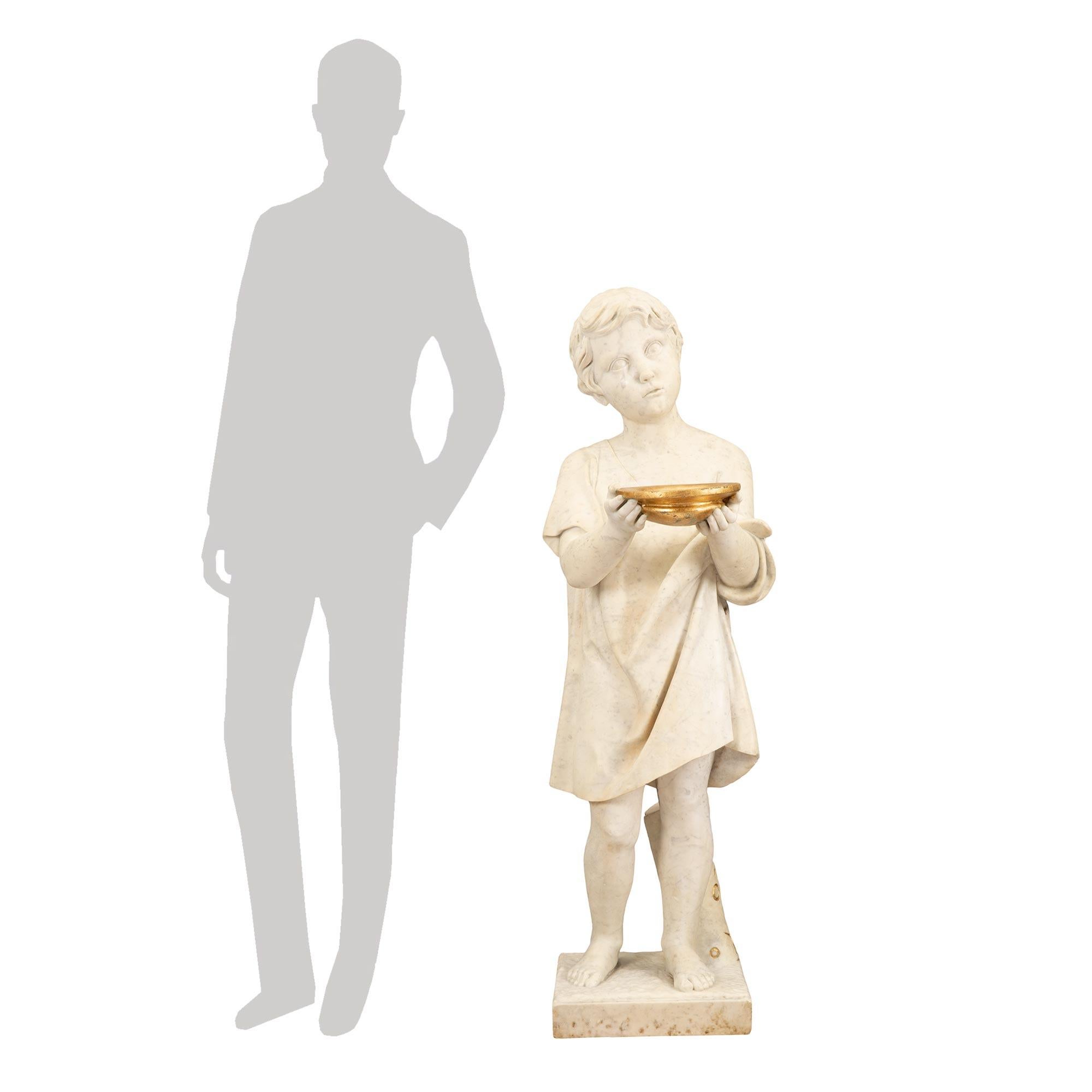 A most charming Italian 19th century white Carrara marble statue of a young boy with gilt accents. Raised by a rectangular base with a terrain design and a tree trunk, with fanciful carved designs and gilt accents. The young boy is draped in a