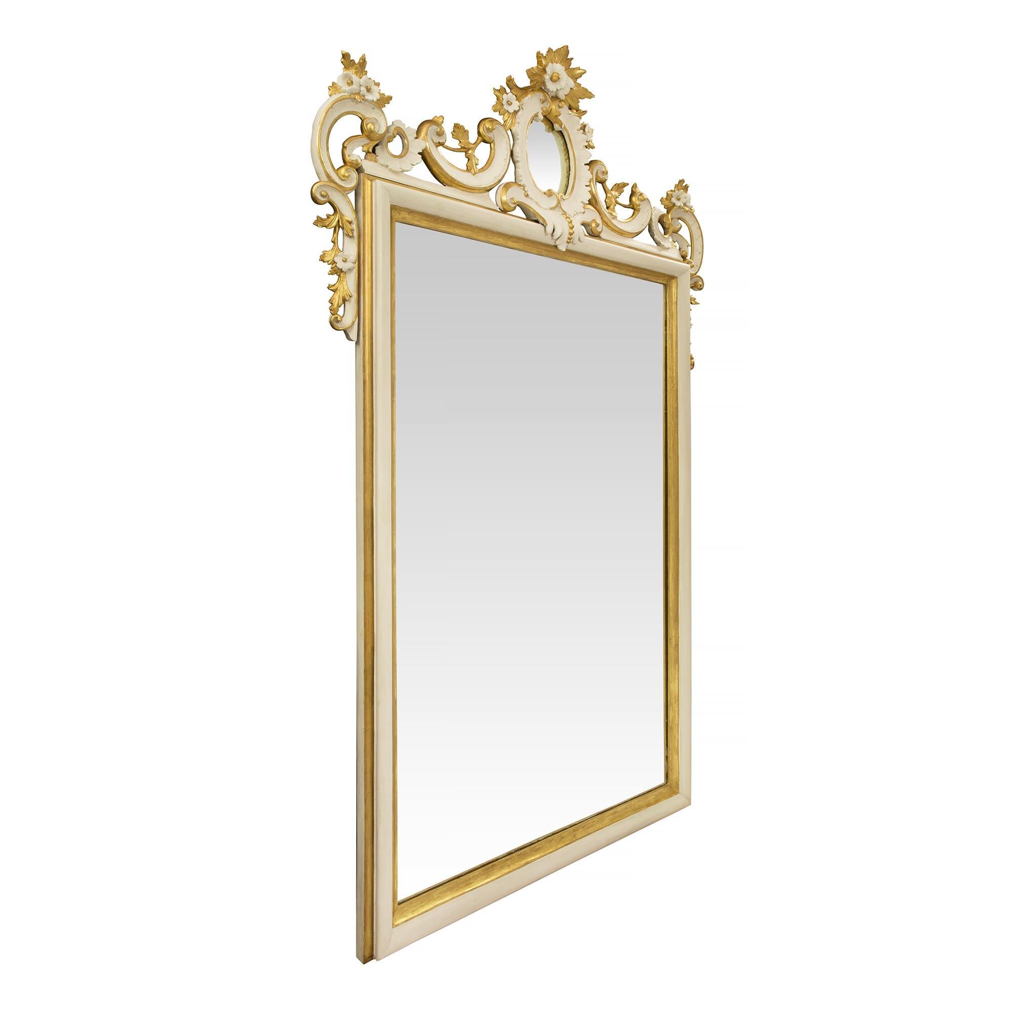Un beau miroir italien du 19ème siècle en bois blanc polychrome et doré. La plaque de miroir est encadrée d'une fine bordure rectangulaire polychrome blanche et mouchetée de bois doré. La saisissante couronne supérieure percée est centrée par une