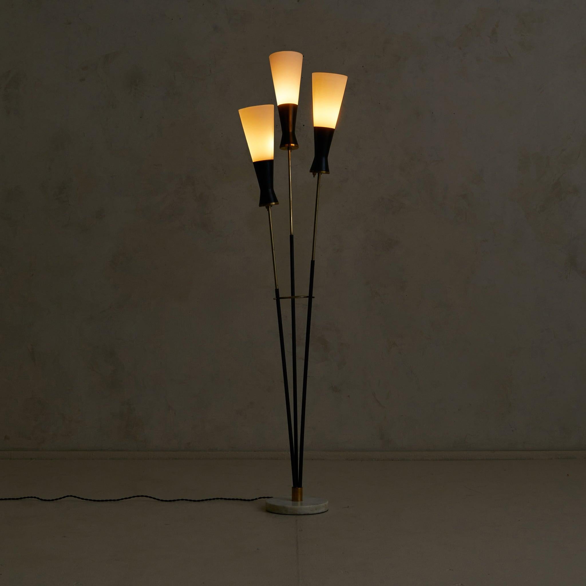 An Italian floor lamp dating to the 1950s. Three slender stems rise in rhythmic offset, their black-lacquered metal bodies cinched with brass fittings and joined by a circular brass ring. Each arm holds a flared opaline glass shade—softly tapering