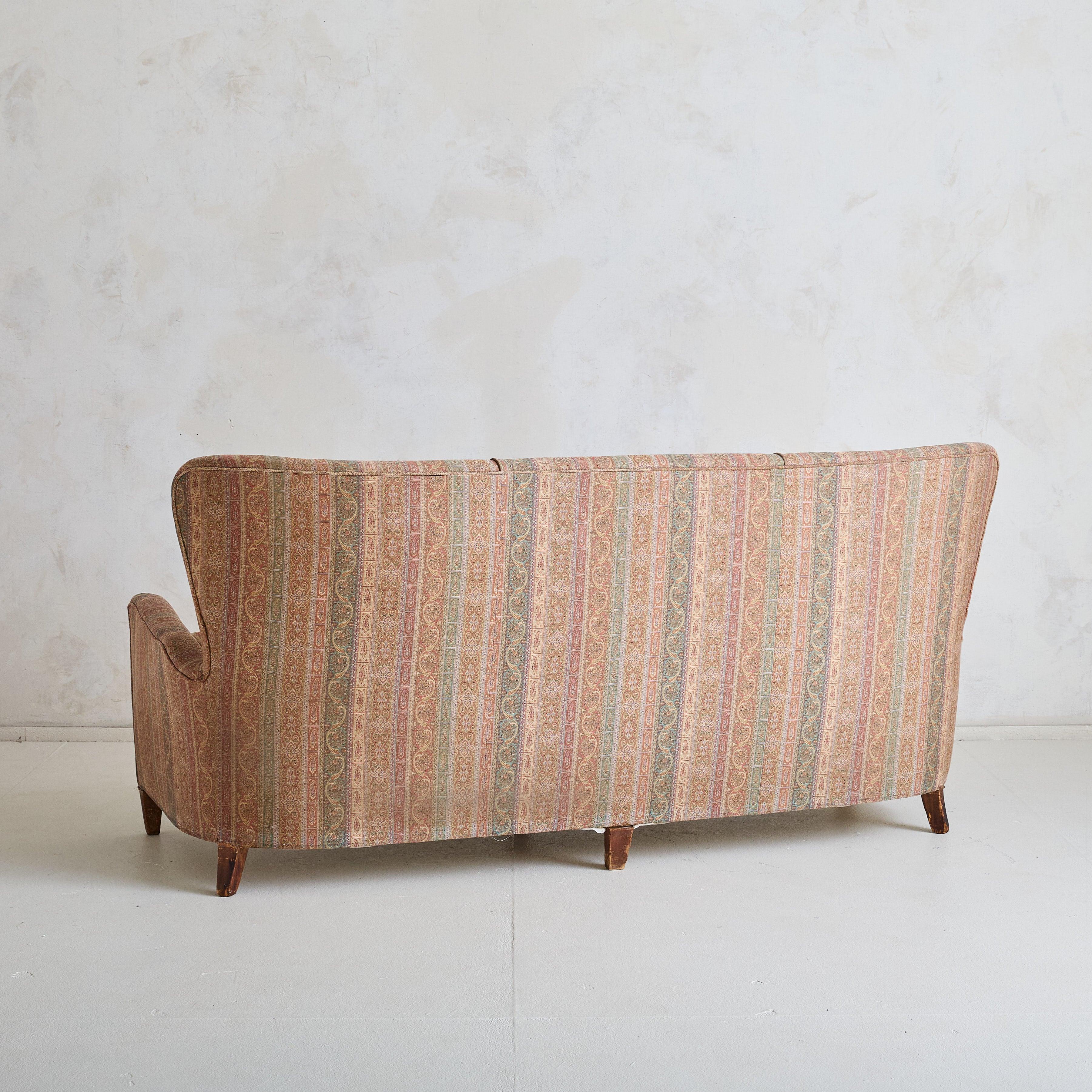 This Italian three-seat sofa from the 1960s epitomizes mid-century design with its soft, sculptural form. The high, button-tufted backrest and gently sloped arms define the silhouette. Tapered wooden legs provide a light, airy lift, balancing its