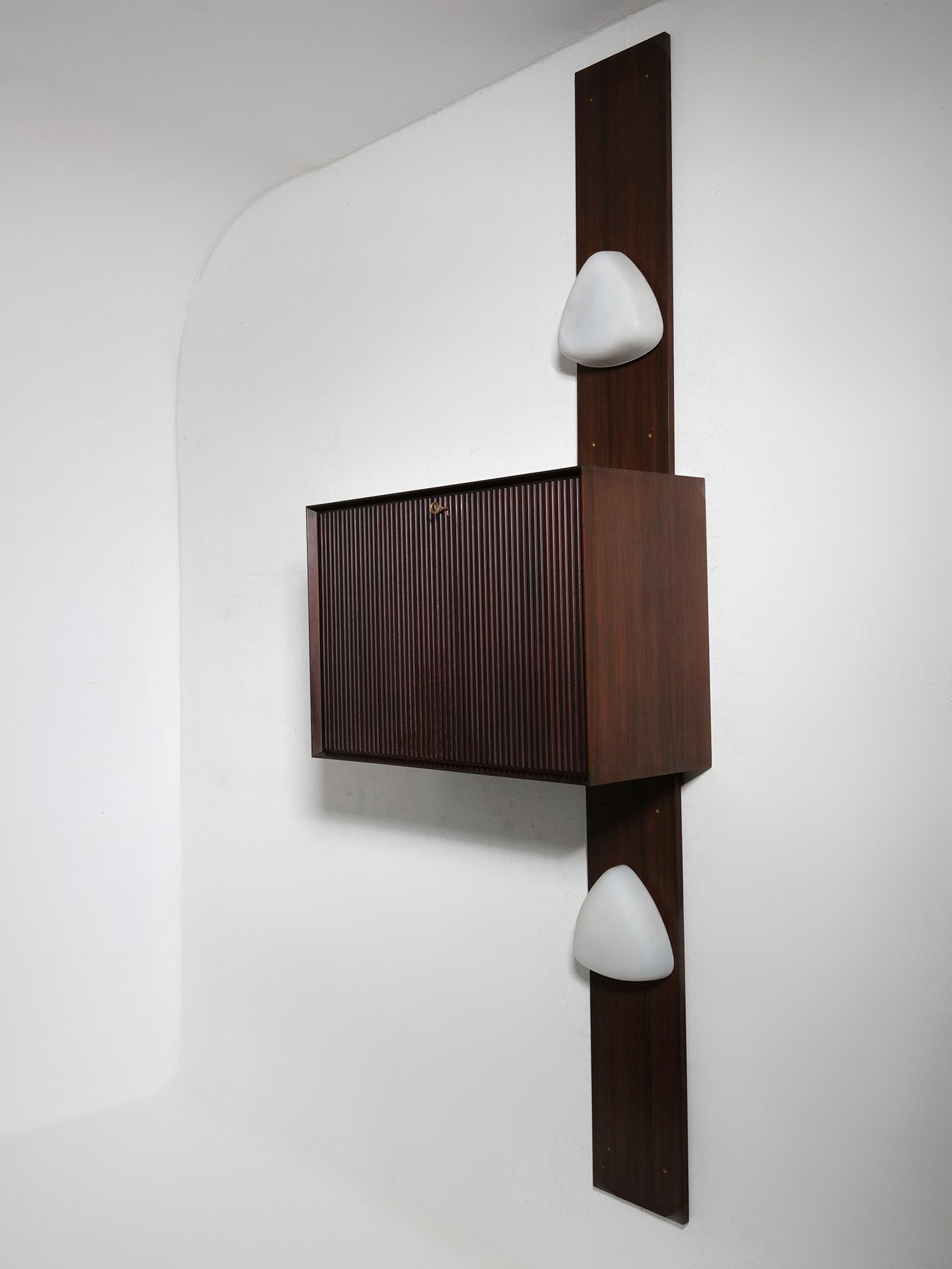 Wall-mounted dry bar.
Central unit feature a flap door and mirrored inside with glass shelves.
Main piece is surrounded by two vertical wood panels with opaline glass sconces.