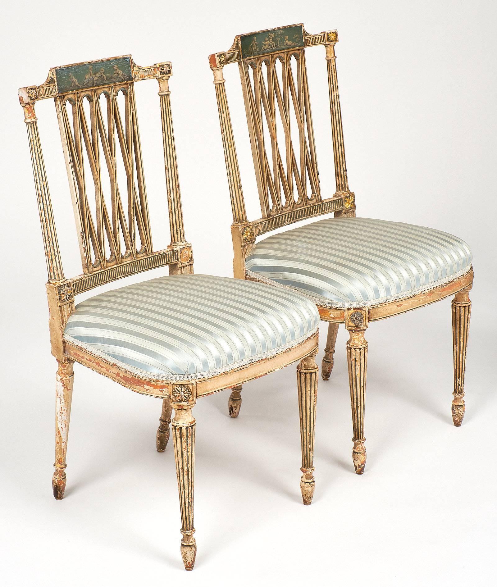 Pair of antique Italian painted side chairs. Crests are painted in blue and green with four grisailie painted putti on each chair in classical motifs above four “x” forms defining the back between reeded stiles. The chairs are upholstered in a light