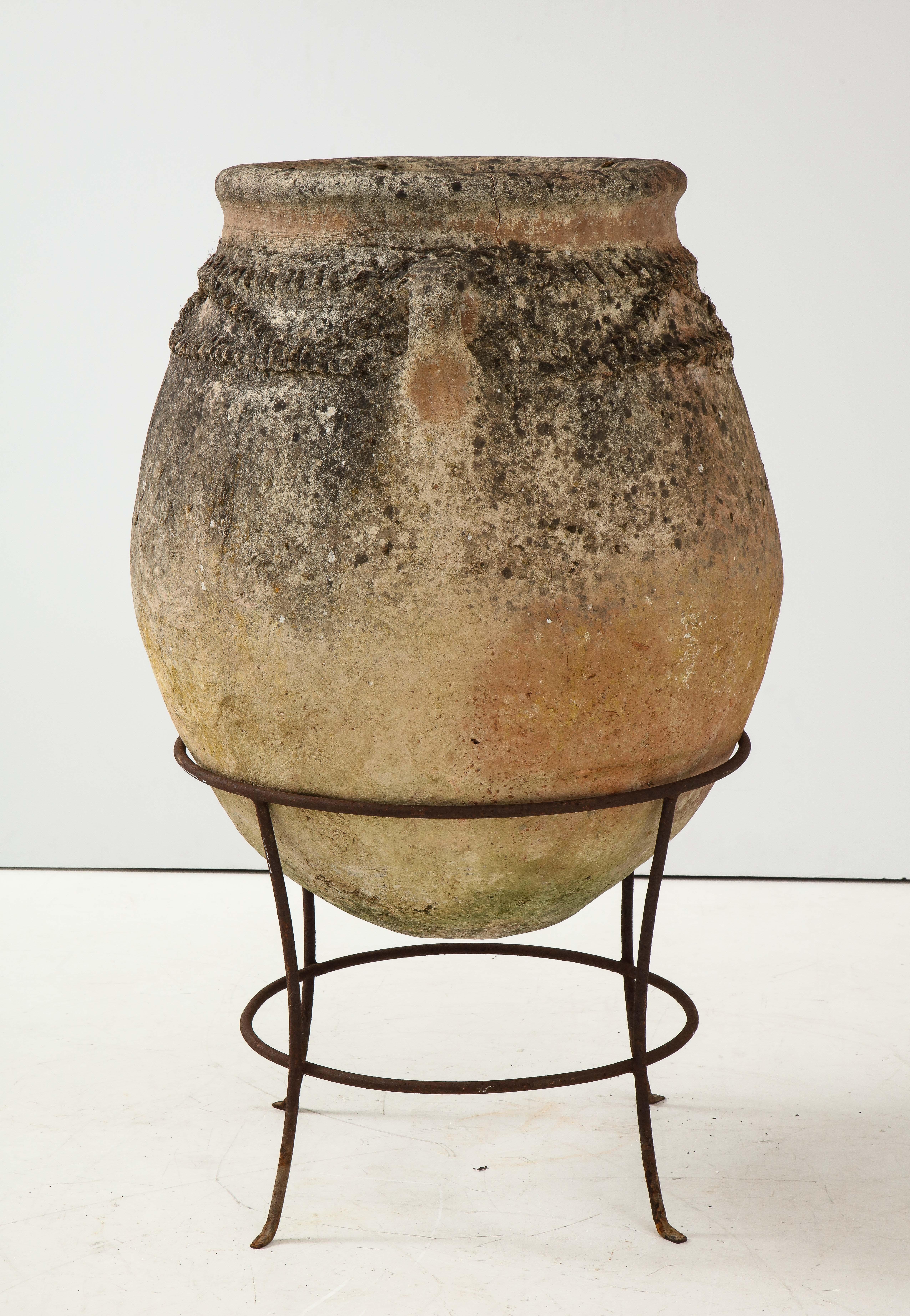 An Italian antique terracotta olive oil jar, of very grand scale and mounted within its original iron stand. The jar has moss and beautifully aged patina with two handles and horizonal cross hatch design. A beautiful, organic and dramatic piece for
