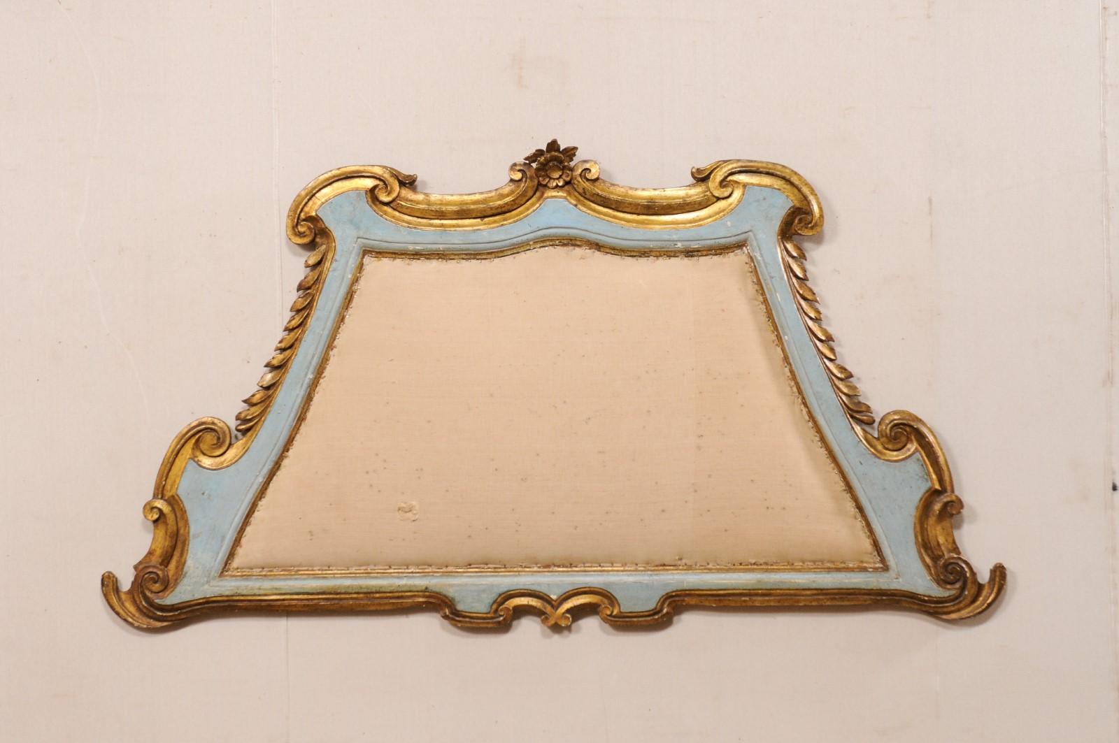 An Italian carved and gilt wall-mounted headboard, with upholstered center, from the early 20th century. This antique wall-plaque from Italy has a trapezium shaped hand carved wood frame, outlined in beautiful c-scrolls, leaves, and a single flower
