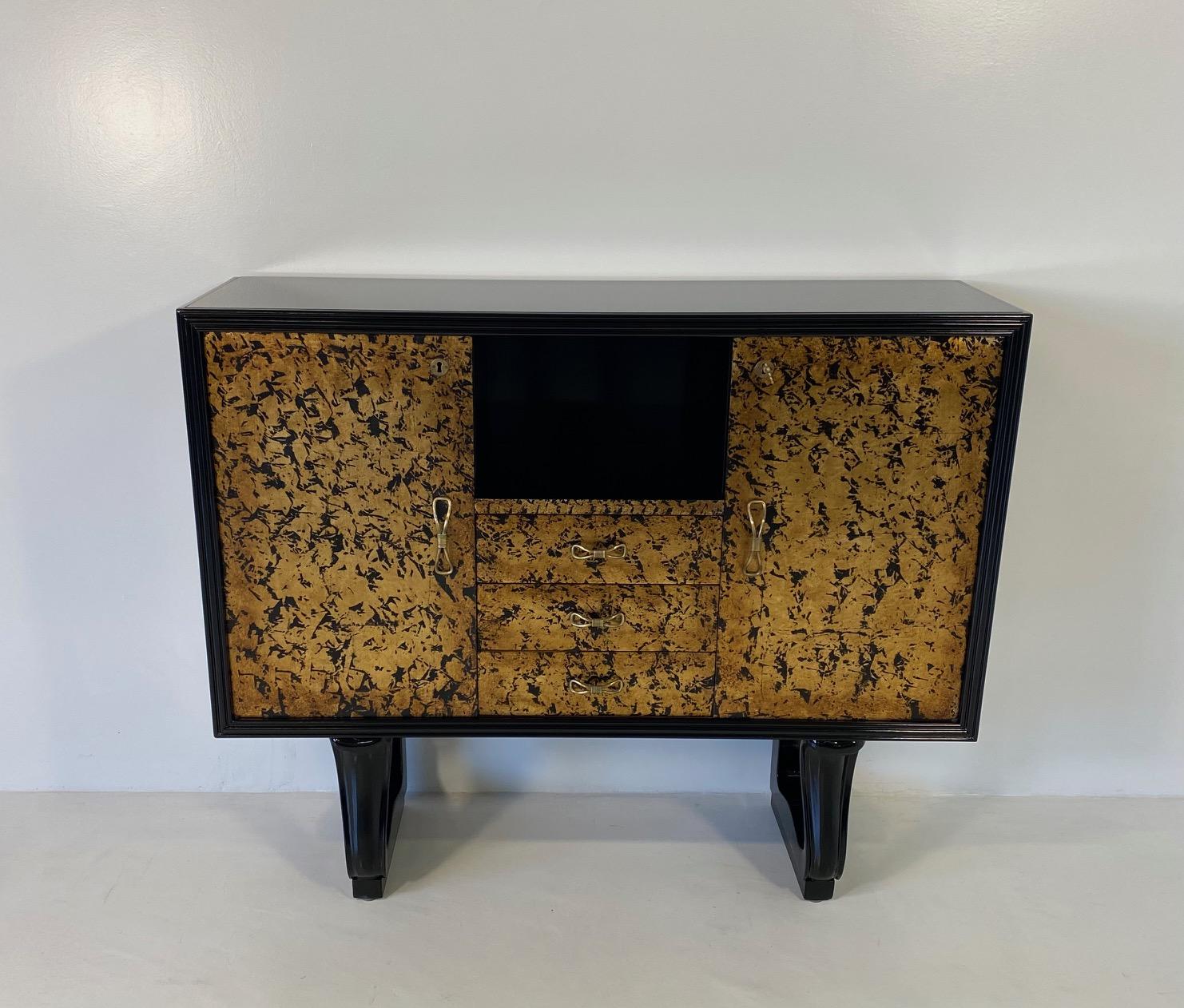 This cabinet was produced in Italy in the 1940s.
The structure is black lacquered, while the three central drawers and the two doors are decorated with gold leaf, the top is a black glass.