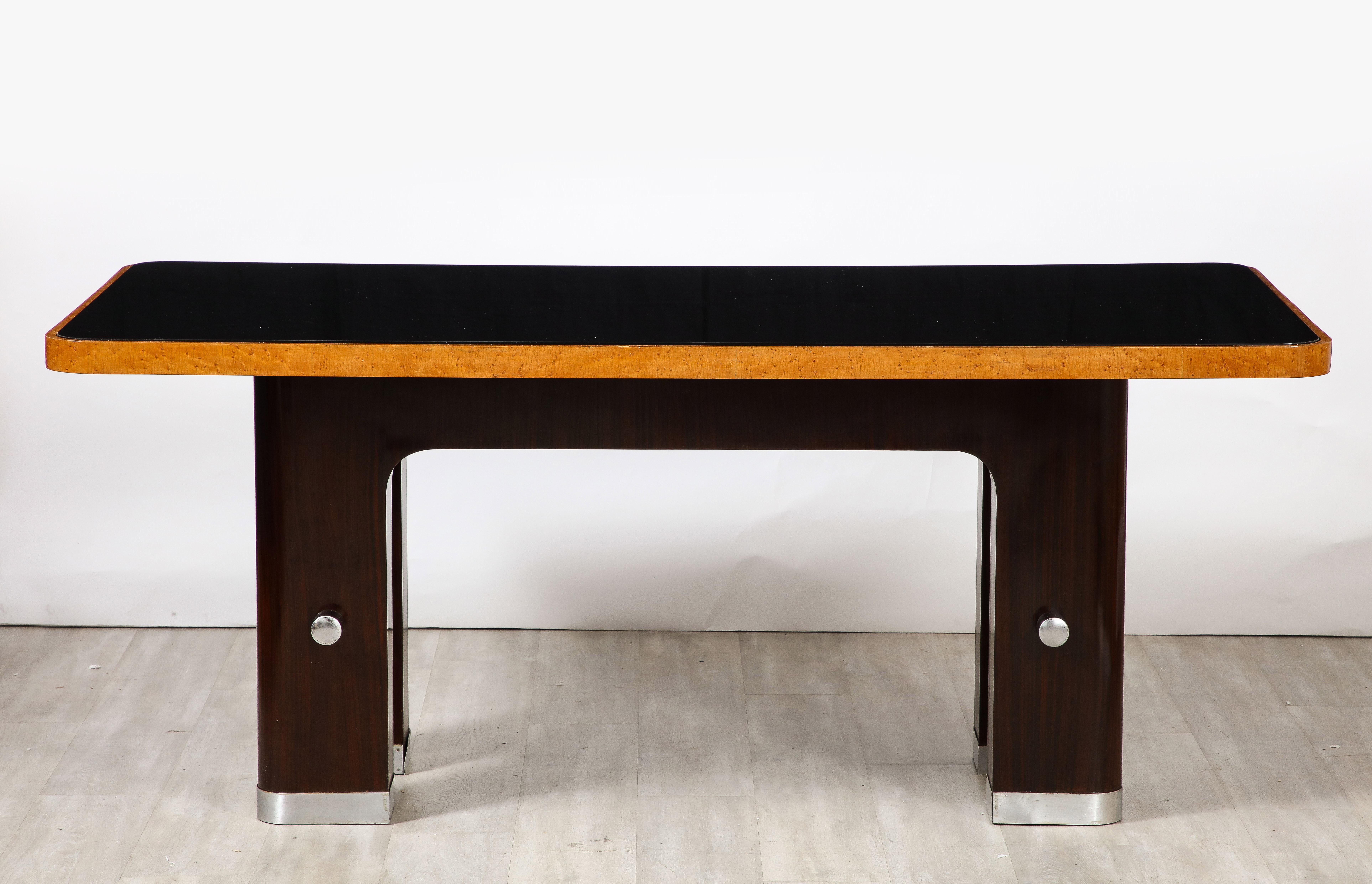 An Italian Art Deco dining table, the black glass surface is inset to the molded apron of Birdseye maple; resting on two rounded Macassar ebony columns on each end connected with a rounded chrome stretcher, the chrome echoed at the base of each