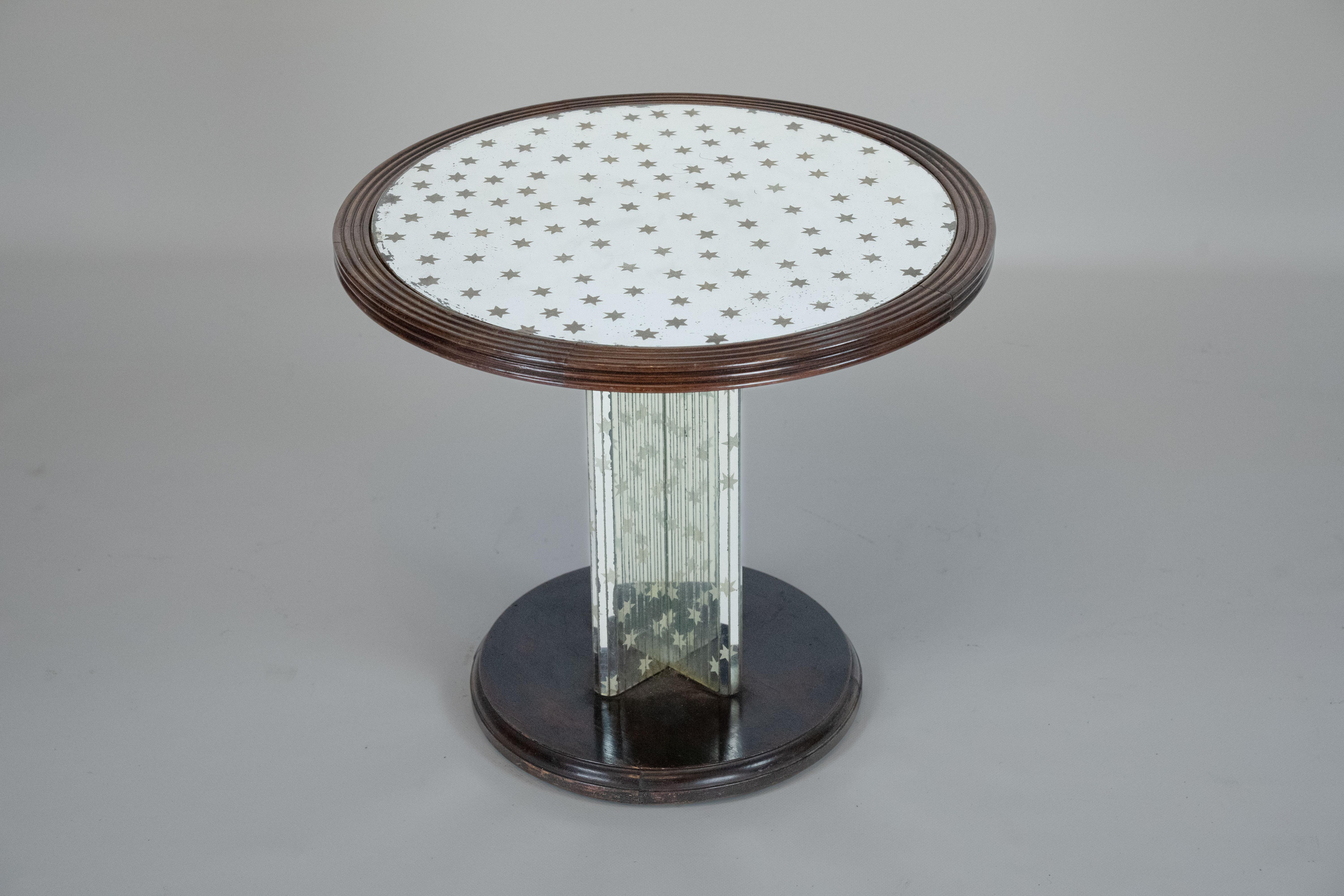 The table features a round wooden top with a mirrored surface decorated with an elegant pattern of small stars. Its central pedestal is composed of mirrored panels, creating a refined play of reflections and light. The base is crafted in dark wood,