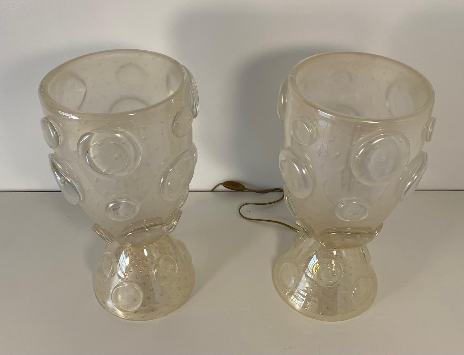 Cette paire de lampes vases a été produite en Italie, à Murano, la capitale mondiale de la verrerie. 
Elles sont entièrement réalisées en verre blanc de Murano, avec des bulles d'or à l'intérieur. Les cercles décoratifs sont également fabriqués