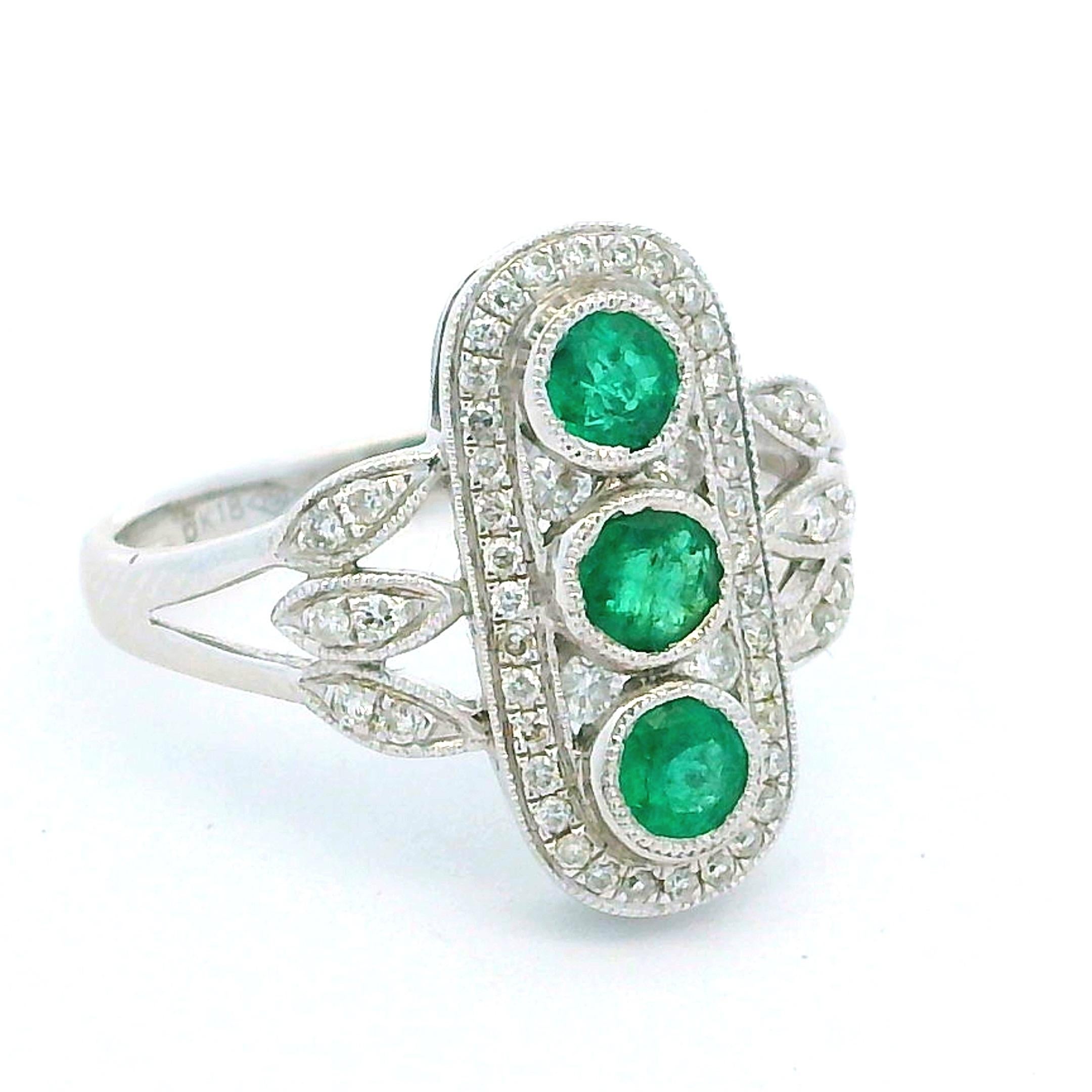 Cette bague italienne de style Art déco, d'un grand raffinement, met en valeur trois émeraudes rondes vibrantes disposées verticalement dans un panneau classique allongé. Les émeraudes (environ 0,55 carats au total) sont encadrées par un délicat